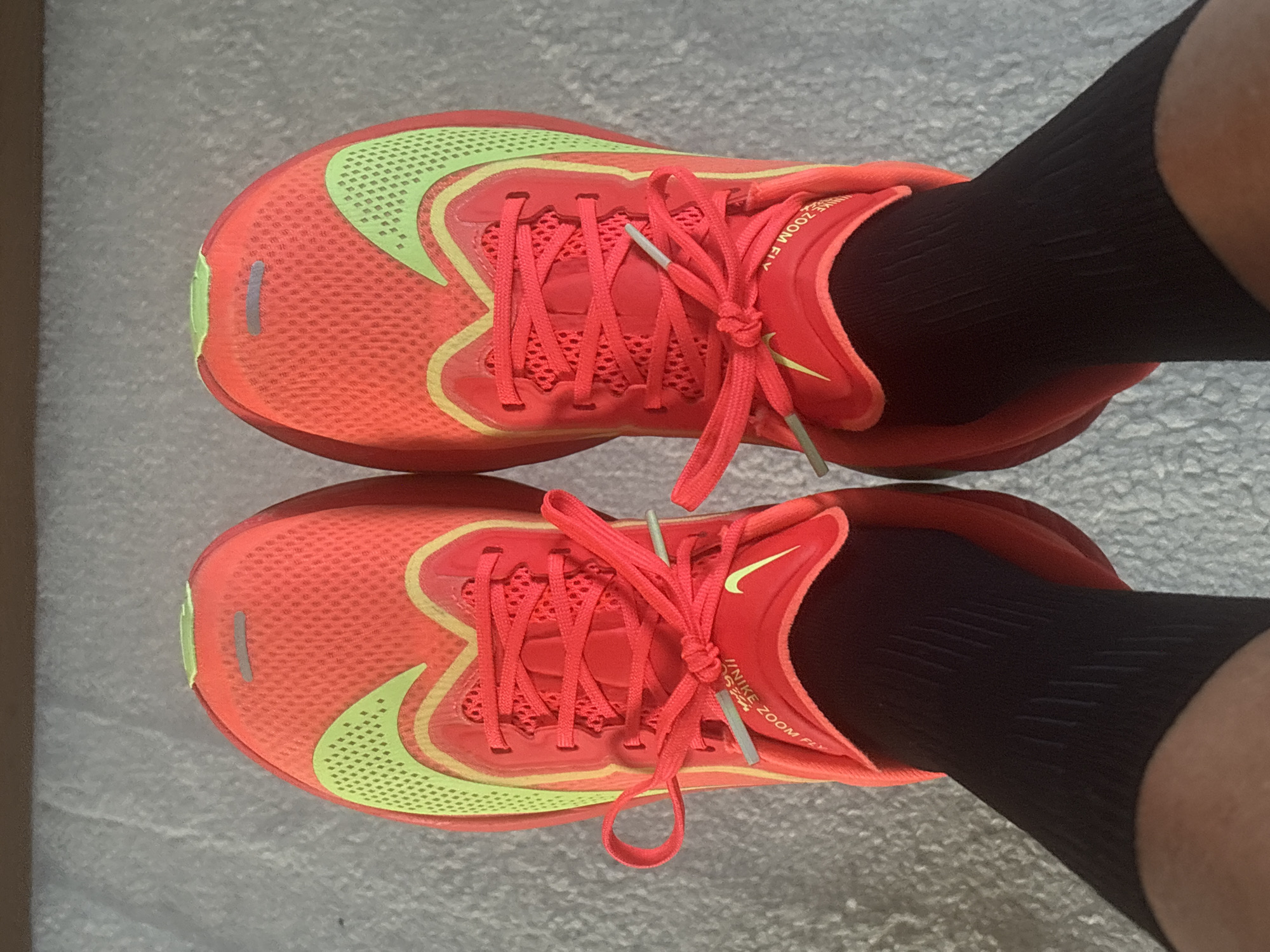 An overhead shot captures a person's feet, clad in black socks and bright running shoes, positioned on a light grey, textured floor, possibly indoors in Pétange, Luxembourg.
The two shoes are identical, featuring a vibrant coral-red or orange-red main color with distinctive lime-green accents. A large Nike swoosh is prominently displayed on the side of each shoe in the same lime-green. A thinner, lighter yellow-green line outlines various sections of the shoe's upper, including parts of the swoosh and eyelets. The shoes are laced with matching coral-red laces, tied in neat bows, and feature silver-grey aglets (lace tips).
Visible text on the side of the shoe collar clearly reads "NIKE ZOOM FLY", indicating the model. The shoe's upper material appears to be a breathable mesh. A small, vertical, reflective grey stripe is present on the toe box of each shoe, along with a subtle lime-green trim at the very front tip. The person's black socks rise above the shoe collar and show a ribbed or textured pattern. The overall impression is one of modern athletic footwear.