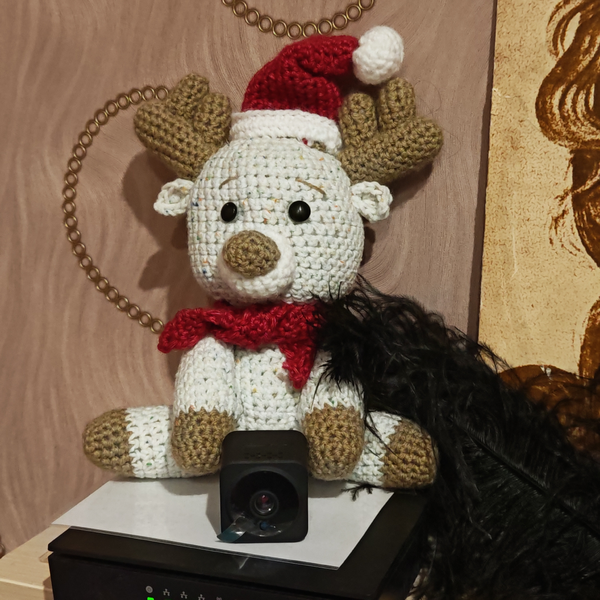 The image displays a festive indoor arrangement featuring a crocheted reindeer plush toy. The reindeer, predominantly white with subtle multi-colored flecks, has brown crocheted antlers and snout, black button eyes, and white ears. It is adorned with a red Santa hat, complete with a white brim and pom-pom, and a red crocheted scarf around its neck.
The reindeer is seated on a white sheet of paper, which in turn rests on a black electronic device that shows several illuminated green indicator lights. In front of the reindeer, a small, black, square-shaped camera or surveillance device is positioned, with its lens visible and a small piece of blue tape adhered to its front. To the reindeer's right, a large, fluffy black feather boa or decoration is draped.
The background consists of a textured light brown wall. On the left side of the wall, a decorative golden chain-link pattern is visible, curving around the reindeer's head. On the right, a section of a beige and brown abstract artwork or wallpaper with flowing, organic patterns can be seen. The overall scene suggests a holiday or festive decorative display. No discernible text is visible within the image. The setting is an indoor room in Kladovitsy, Russia.