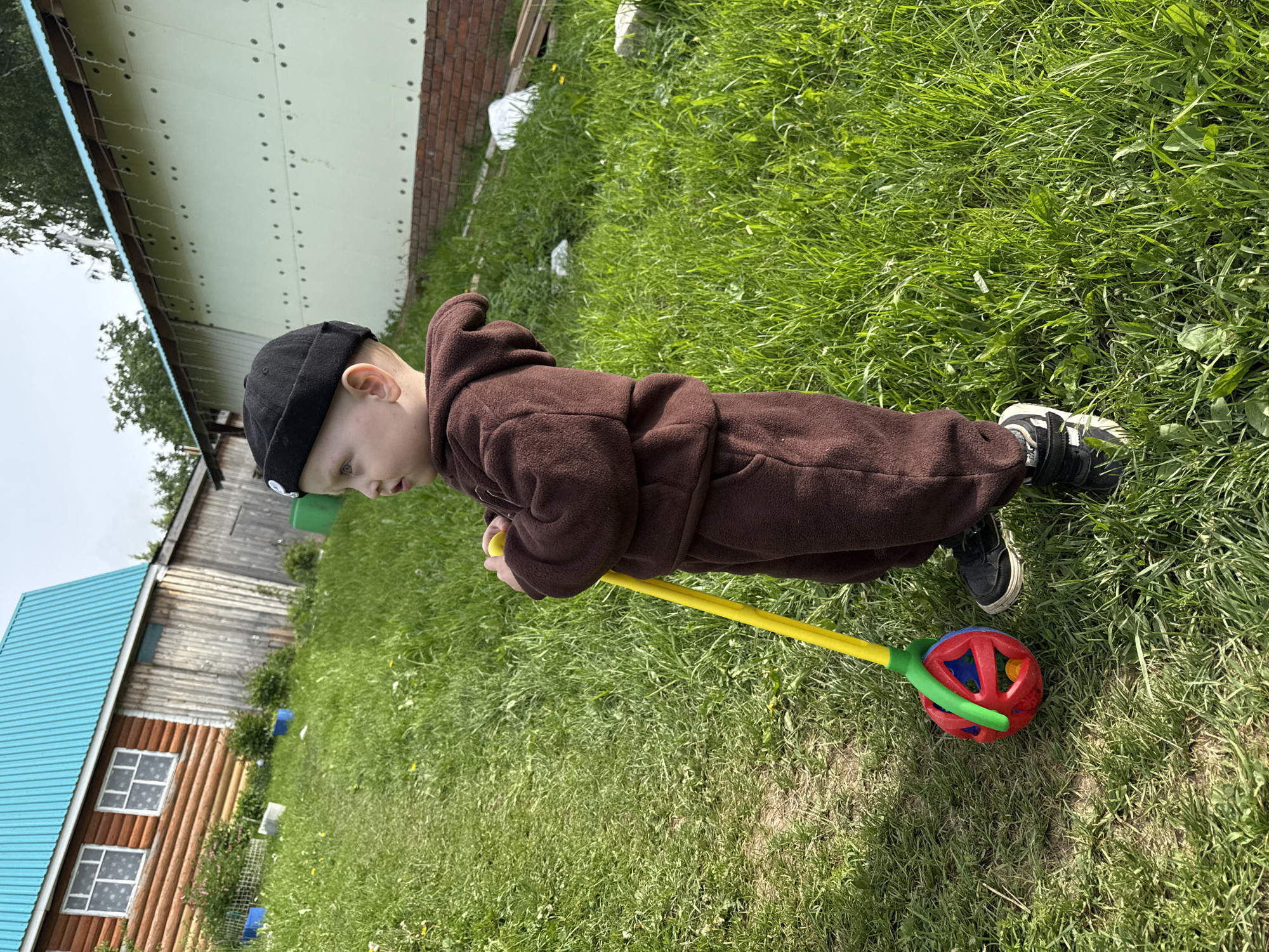 A young child, likely a toddler, is depicted outdoors in what appears to be a rural or backyard setting. The child is dressed in a dark brown hooded jumpsuit or overall, a black beanie, and black and white sneakers. They are holding and pushing a yellow stick toy with a colorful red, blue, and green ball-like mechanism at its end, which is resting on the grassy ground. The child's posture and focused gaze indicate active engagement in play.
The scene is dominated by bright green grass underfoot. In the background, there are several buildings. To the left, a wooden structure with a vibrant blue corrugated metal roof and white-framed windows is visible. Further back and to the right, another building features a light green or white metallic-clad wall with visible rivets, and a brick foundation. The overall impression is a bright daytime scene, suggesting sunny or partly cloudy weather, with clear shadows. There is no visible text in the image. The location is contextualized as Ляльшур, Russia.