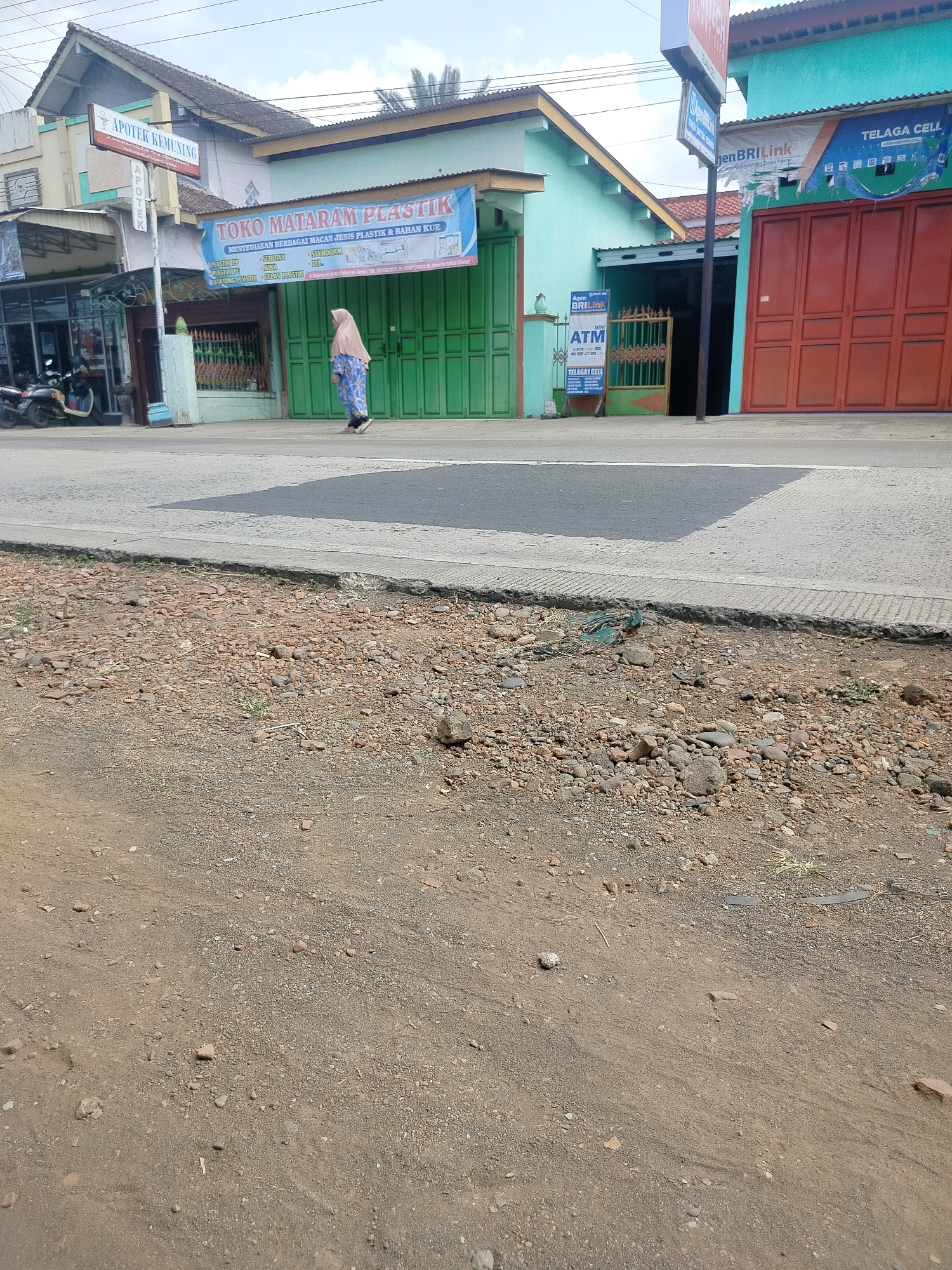 The image depicts a street scene in Kroya, Indonesia, with a focus on storefronts and the surrounding environment. In the foreground, a dirt and gravel roadside is visible, with scattered small stones and patches of sparse vegetation. A concrete curb separates the road from the sidewalk.
On the sidewalk, several buildings line the street. To the left, a building with a light-colored facade features a sign that reads "APOTEK KEMUNING" (Kemuning Pharmacy). Below this, another sign in white text on a dark background says "APOTEK" (Pharmacy). A motorcycle is parked in front of a darkened storefront.
Adjacent to the pharmacy, a business with a blue banner is labeled "TOKO MATARAM PLASTIK" (Mataram Plastic Shop), advertising the sale of various plastic items and household goods. The shop has green double doors.
Further to the right, a mint green building has a doorway and a smaller green gate. A sign for "Agen BRIlink" (BRIlink Agent) and "MINI ATM" is posted near the gate. Another sign for "TELAGI CELL" is visible on the facade of a building with red double doors.
A woman wearing a light-colored headscarf and a patterned blue and white outfit is walking on the sidewalk, moving away from the viewer. The sky above is overcast. The scene appears to be in a commercial area of the city.