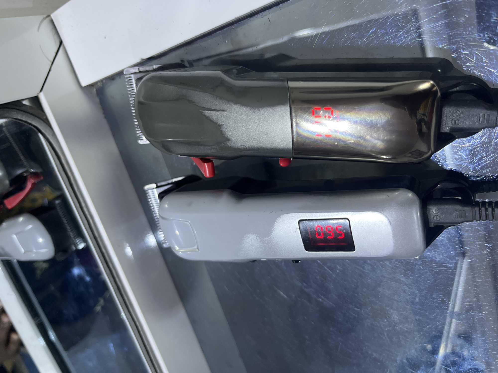 Two hair clippers are positioned on a reflective surface. The upper clipper is gray with a red toggle and a digital display showing "09." The lower clipper is light gray with a digital display showing "095." Both clippers are plugged in. The image's context is unknown, and no information suggests its location is Gura Top, Nigeria.