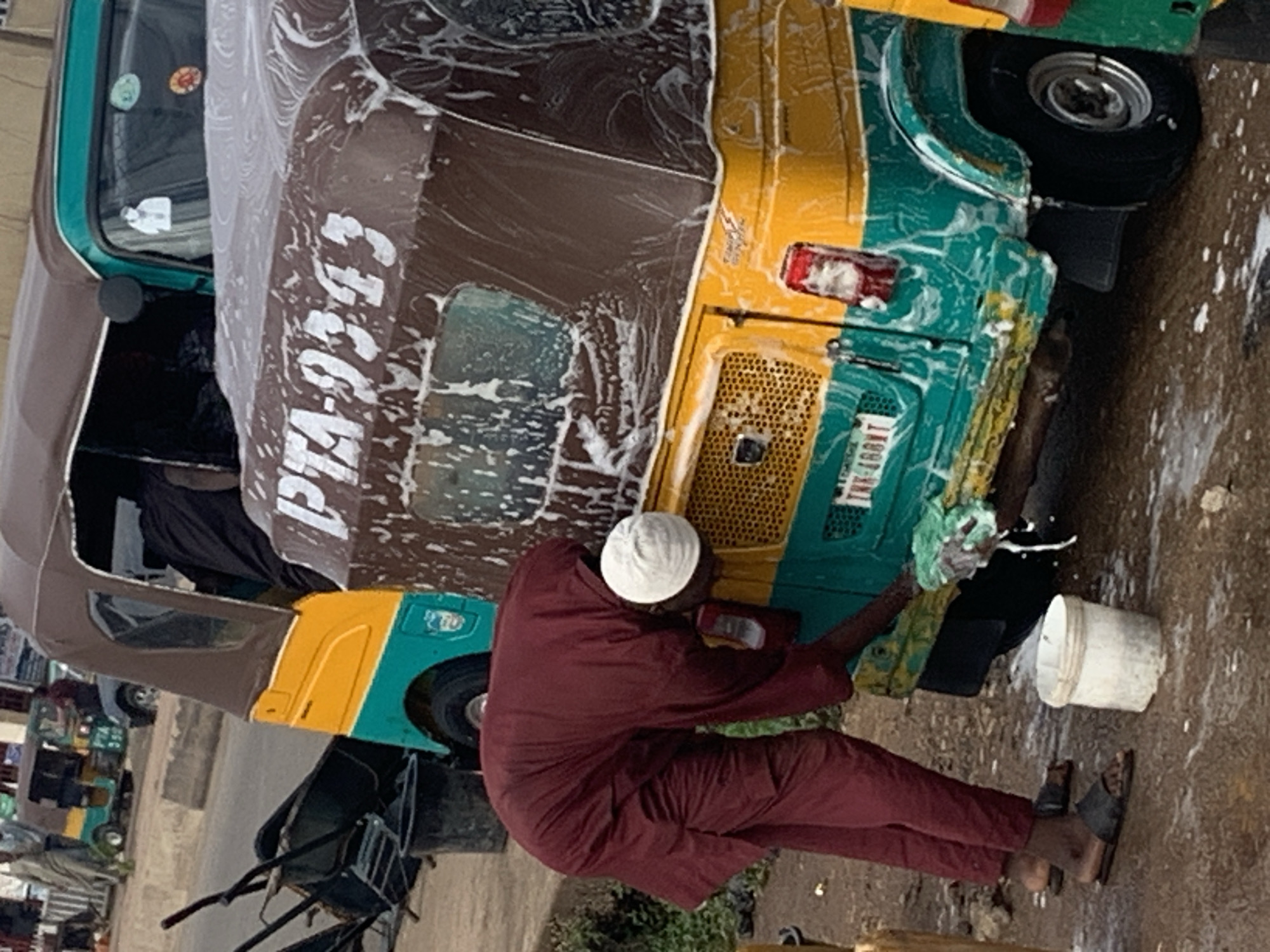 The image depicts a street scene in Gura Top, Nigeria, where a person is washing a three-wheeled vehicle, commonly known as an auto-rickshaw or Keke Napep. The auto-rickshaw is predominantly brown and yellow with a teal lower body. White soap suds cover a significant portion of the vehicle, indicating the washing process is underway. The person, dressed in a maroon outfit and a white cap, is bent over, actively scrubbing the vehicle with a green sponge. A white bucket is visible on the ground near the person's feet, likely containing water and cleaning supplies. In the background, other vehicles and structures suggest a typical urban environment. The ground appears to be wet and dirty, consistent with a washing area on a street. A license plate on the rear of the vehicle reads "PTA-9343".