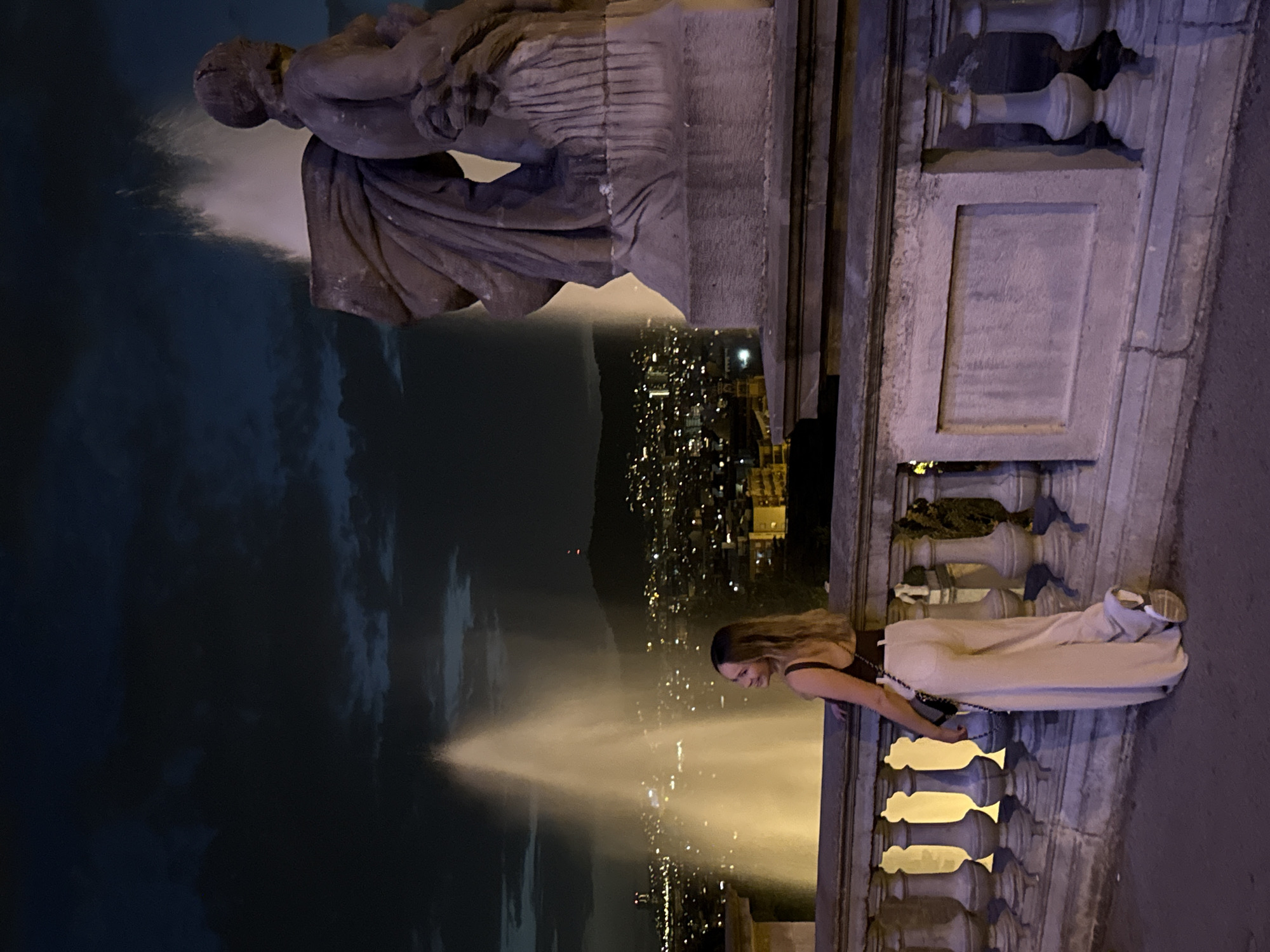 In Barcelona, Spain, at night, a woman stands on a balcony overlooking a city illuminated by lights. Two large fountains spray water into the dark sky. A stone statue of two figures is visible on the right side of the frame. The woman is wearing a dark top, light-colored pants, and white sneakers. She is looking to her left, with her right hand resting on the railing. A black purse with a chain strap is slung over her left shoulder. The railing is made of stone and has decorative balusters. The city lights form a glowing panorama in the background, with hills visible in the distance. The sky is dark with some clouds.