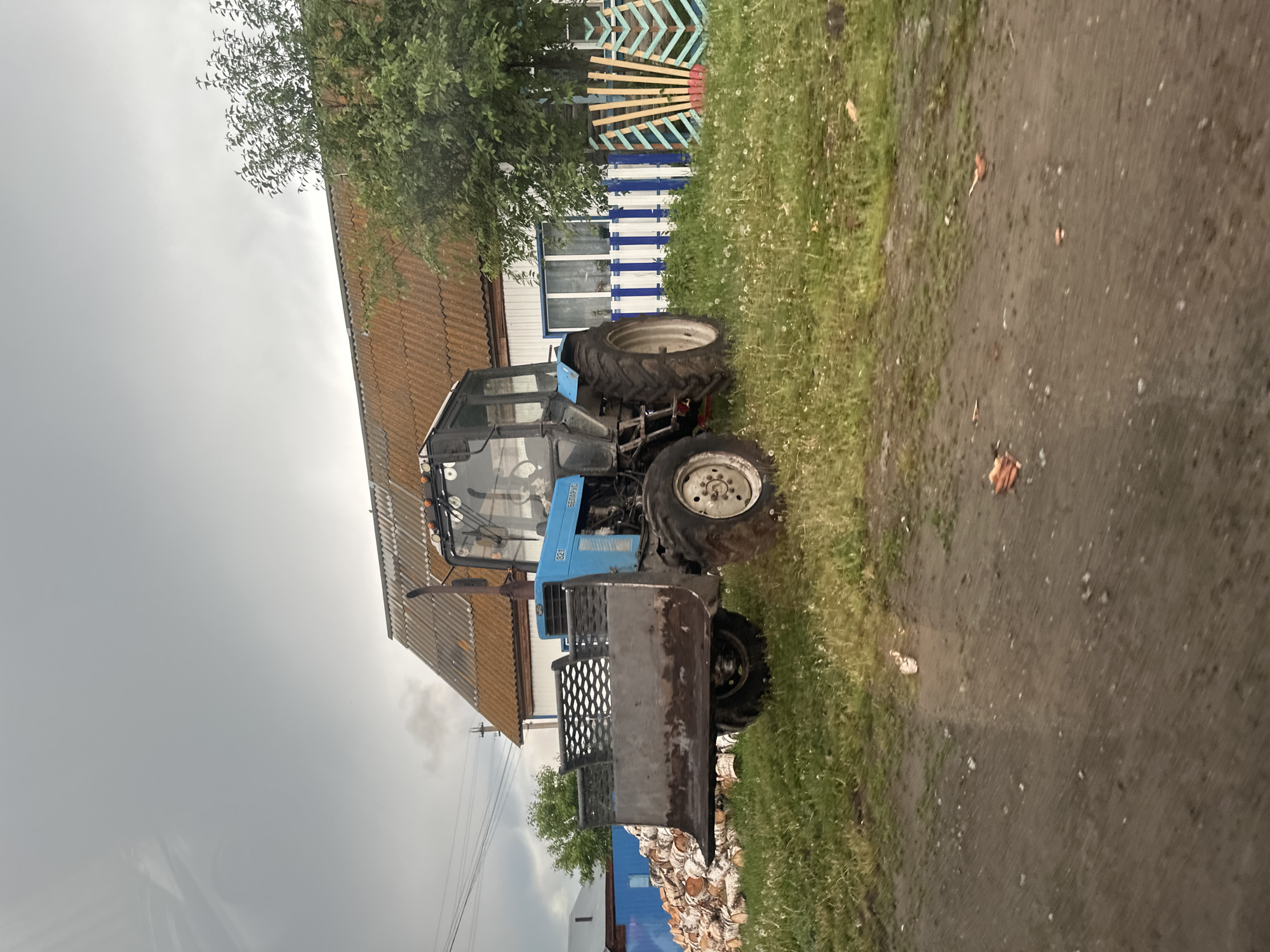 A blue MTZ-821 "BELARUS" tractor, equipped with a front-mounted loader/blade attachment exhibiting rust along its lower edge, is parked on a grassy verge in an outdoor rural or village setting. The tractor faces slightly right, with its left side and rear wheel visible.
Behind the tractor, a single-story house with white walls and a reddish-brown corrugated roof is visible. A prominent exhaust pipe from the tractor extends upwards in front of the house. To the right of the tractor, a wooden picket fence with alternating white and blue vertical stripes runs in front of the house, followed by a section of decorative wooden fencing with light blue and natural wood chevron patterns. A leafy tree stands behind this fence section.
To the left of the tractor, a large stack of cut firewood, appearing to be birch logs, is piled against a blue fence or structure. Further left, utility poles with overhead power lines are visible. The foreground consists of a dirt path or road with scattered small stones and several pieces of broken reddish-brown material, possibly terracotta tiles. The grassy area between the path and the house is dotted with numerous white dandelion seed heads. The sky above is uniformly overcast and light grey. No individuals are present in the scene. The location is Unknown, Unknown.