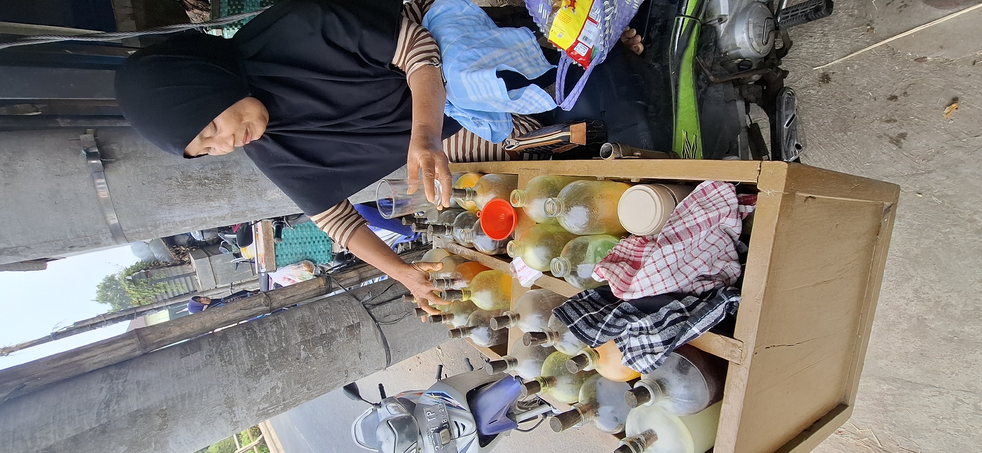 An elderly Indonesian woman wearing a black hijab and striped long-sleeved shirt is standing behind a wooden cart filled with glass bottles containing various colored liquids. She is holding a clear glass in her right hand and reaching out to one of the bottles with her left hand. The cart is positioned next to a motorcycle. The background shows a street scene with trees and buildings. The woman appears to be selling the bottled liquids, possibly a local beverage or fuel, from her mobile cart. The scene is likely a roadside stall in Mranggen, Indonesia, a common sight in many Indonesian towns and cities where vendors utilize motorcycles and carts to sell goods.