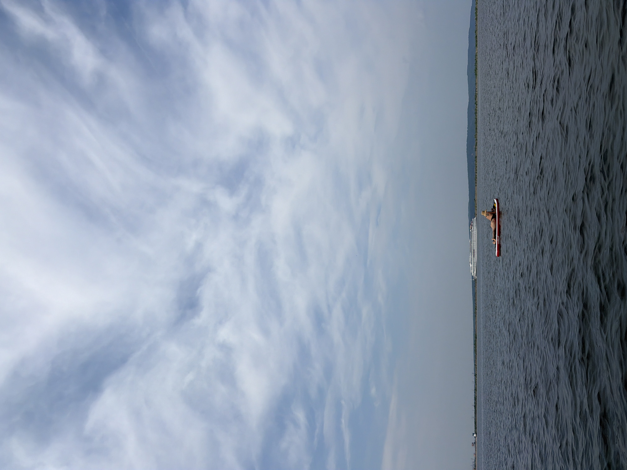 A person is lying on a paddleboard in the Kama River, near Chaikovsky, Russia. The person is wearing a hat and appears to be sunbathing. In the background, a large white cruise ship is visible, as well as the distant shore with trees. The sky is partly cloudy with wispy white clouds. The water is dark and rippled.