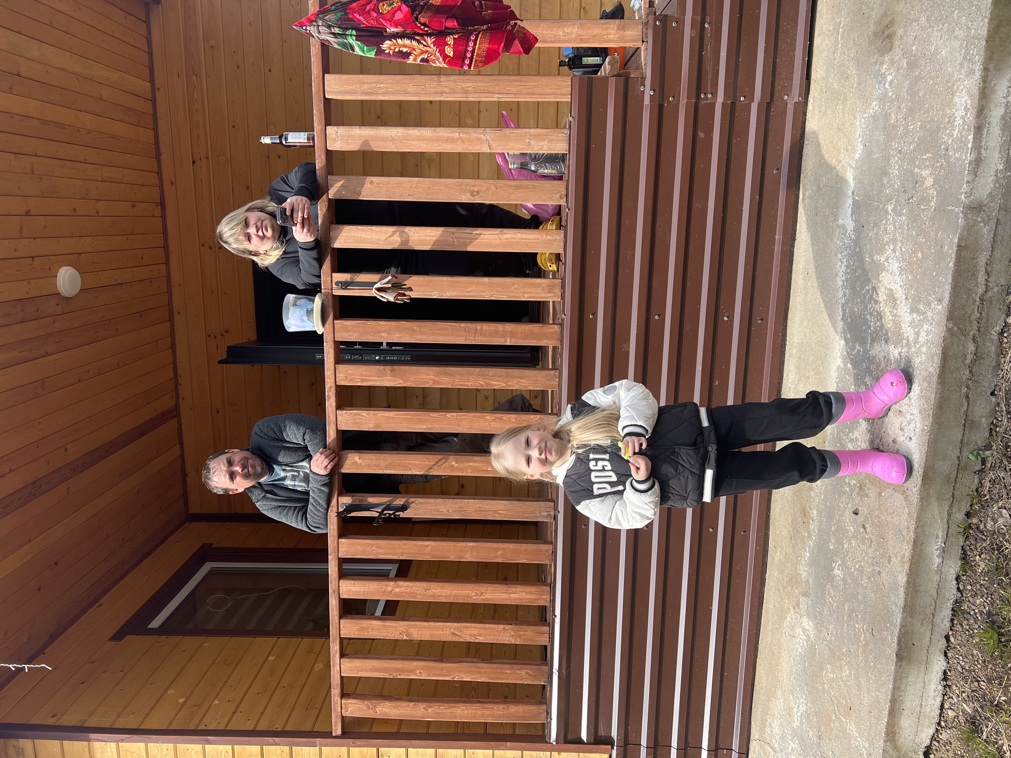 Scene: An outdoor shot featuring three individuals and a child, possibly a family gathering, set against the backdrop of a wooden structure, identified as being in Bol'shakino, Russia.
People:
- Two adults (male and female) are visible on a raised deck, possibly leaning over a railing. The woman is holding a phone.
- A young girl is standing on a concrete surface in front of the structure wearing pink boots.
Objects:
- A building with wood and metal paneling.
- A concrete platform or walkway.
- The woman has a phone.
- The girl is holding something in her hand.
Actions:
- The woman on the deck is holding a phone.
- The girl stands in front of the deck.
Interactions:
- The three people seem to be interacting with each other.
Environment:
- The scene is outdoors, with possible yard or landscaping visible.
- The setting appears to be a residential area or cottage in Bol'shakino, Russia, given the architectural style of the building.