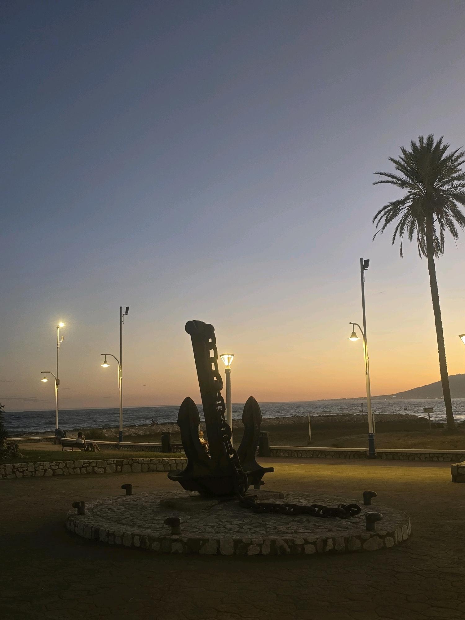 This is a low-angle, full-shot, outdoor, medium-light photograph of a large, dark, metallic anchor with a heavy chain displayed on a circular stone base. The anchor and chain are the central focus of the image. Behind the anchor, a paved walkway leads to a grassy area with a stone wall and a few people sitting on a bench. Beyond the grassy area is the sea, with a faint horizon line and hills in the distance.
The sky transitions from a clear, deep blue at the top of the frame to soft oranges and yellows near the horizon, indicating it is either sunrise or sunset. Several streetlights with warm yellow lights are visible, illuminating parts of the scene. A tall palm tree stands to the right of the frame, its fronds silhouetted against the colorful sky. The scene appears to be a public waterfront area. Given the presence of palm trees, the Mediterranean Sea, and the overall ambiance, this scene is likely located in Málaga, Spain, a coastal city known for its promenades and waterfront attractions.