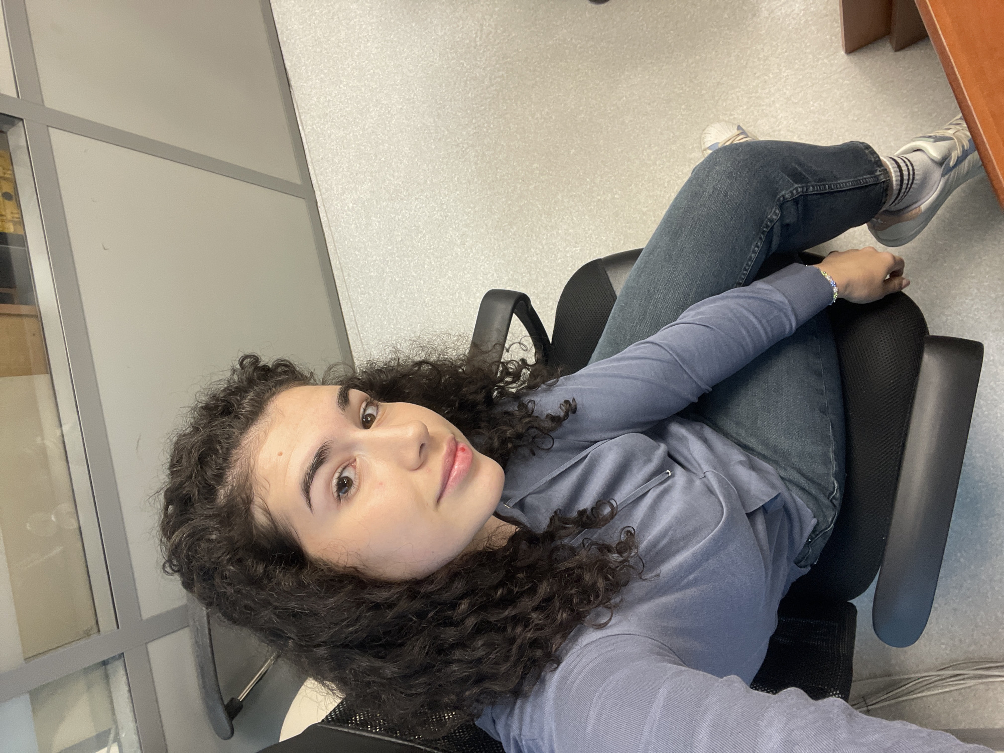 A medium close-up, high-angle selfie captures a young woman sitting in a black office chair within an indoor setting. The woman has dark, curly hair and brown eyes, looking directly at the camera with a neutral, posed expression. She is wearing a light blue-grey long-sleeved top, possibly a hoodie, and dark blue jeans. A subtle, multi-colored beaded bracelet is visible on her left wrist.
Her legs are crossed, and she is wearing white sneakers with light blue or grey accents, paired with white socks that have black stripes. The chair she is sitting in appears to have a black mesh back and armrests. To the right, a part of a light brown wooden desk or table is visible.
The background consists of light grey partition walls or cubicle dividers, framed by grey metal structures, suggesting an office or commercial building environment. The floor is a light grey color with a speckled texture, possibly carpet or textured tile. The lighting is consistent with indoor artificial light. The overall scene indicates an everyday moment in a workspace. No visible text is present in the image. The location context is Sofia, Bulgaria.