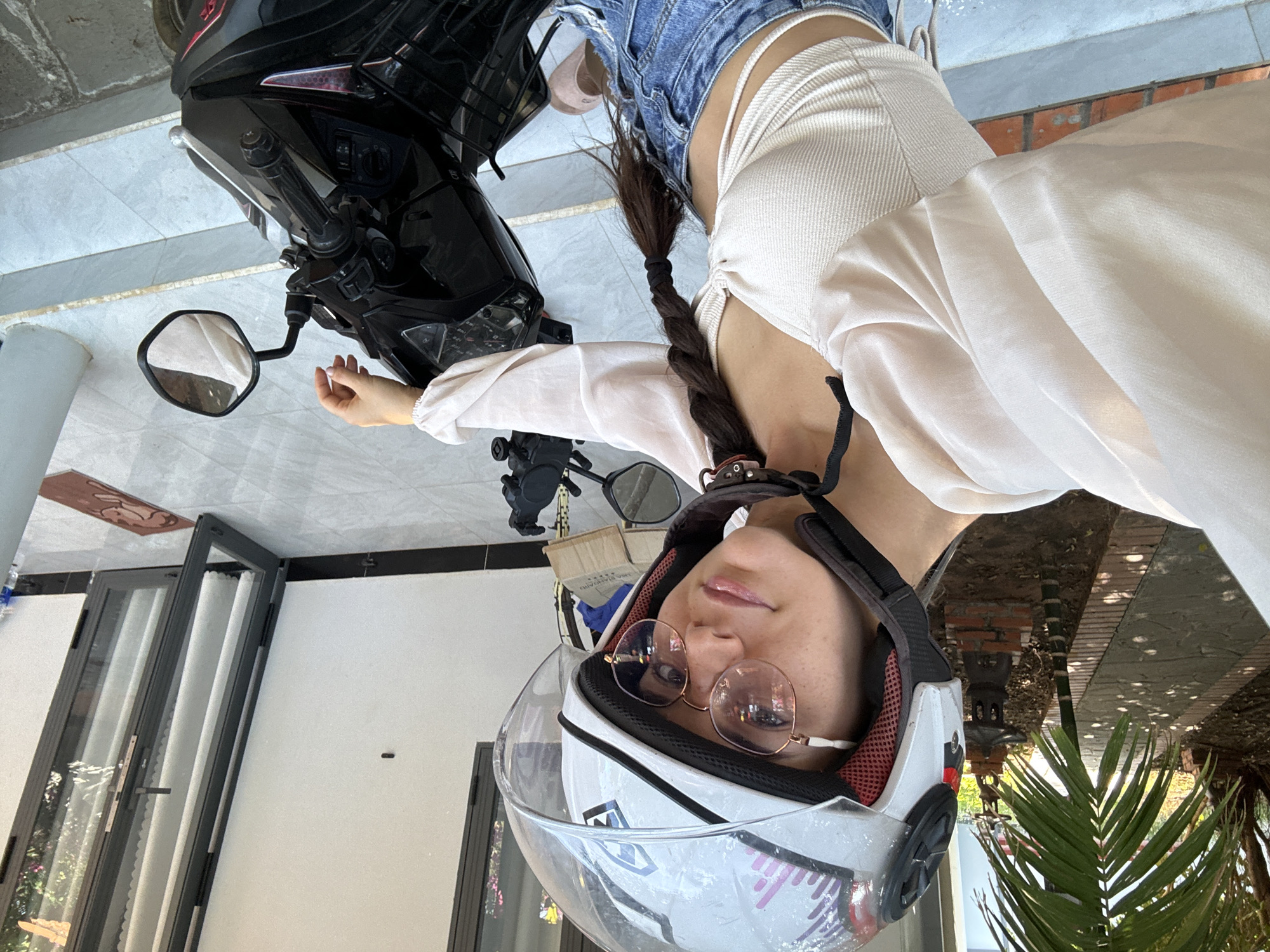 A woman wearing glasses and a motorcycle helmet is indoors, likely in a garage or similar space in Mũi Né, Vietnam. She is wearing a light-colored, long-sleeved top and denim shorts, and her hair is braided. A black motorcycle is partially visible behind her. Palm trees are visible through a door or window.