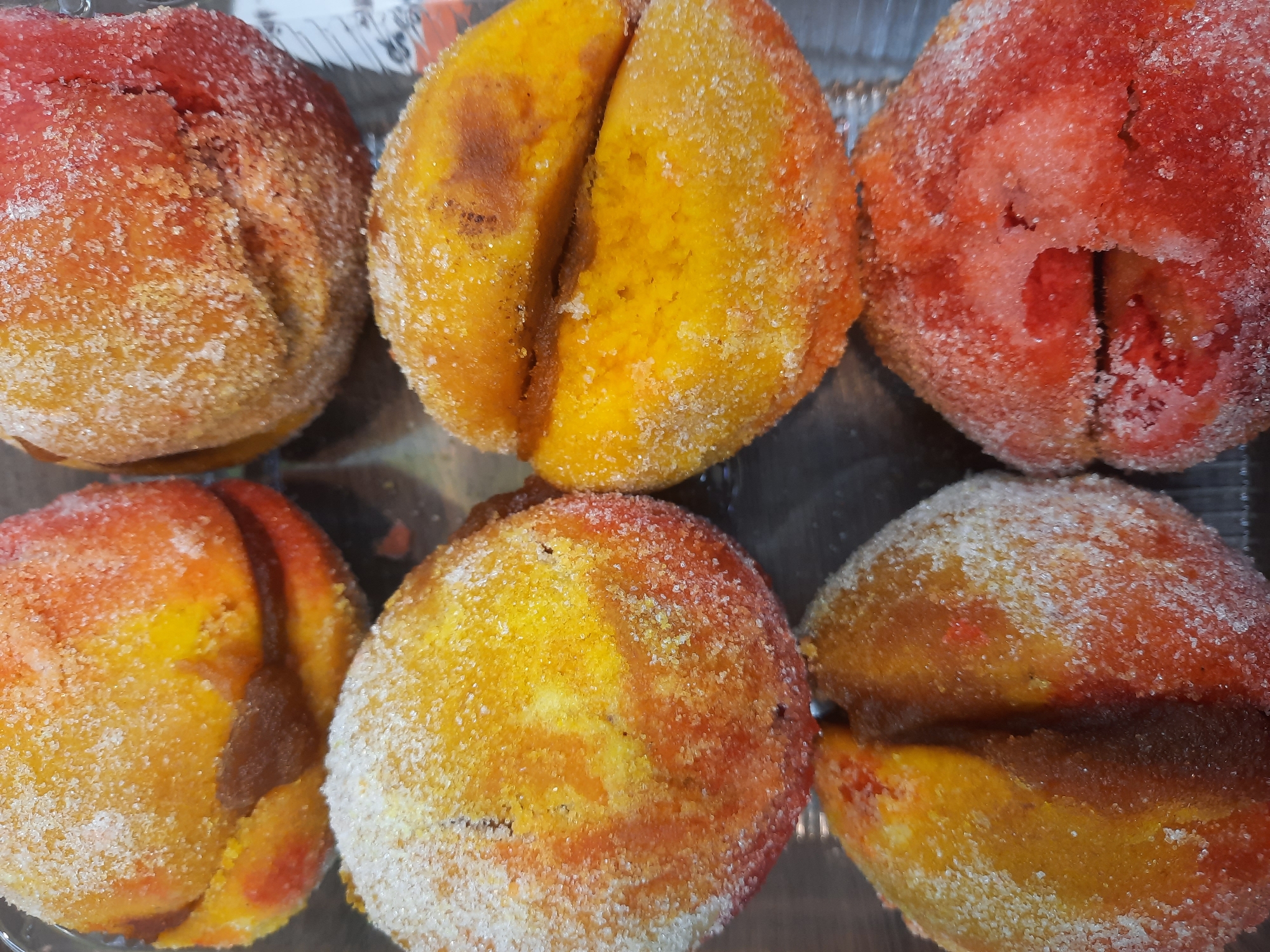 A close-up shot depicts six halved peach-shaped pastries arranged in a clear plastic container. Each pastry is coated with granulated sugar. The pastries are colored in shades of red, orange, and yellow. The fillings are visible in two of the pastries, appearing as dark brown. The pastries are arranged in two rows of three, showcasing their texture and sugar coating. The background is a blurred gray plastic container. The scene is an indoor shot. The location is Unknown, Unknown.