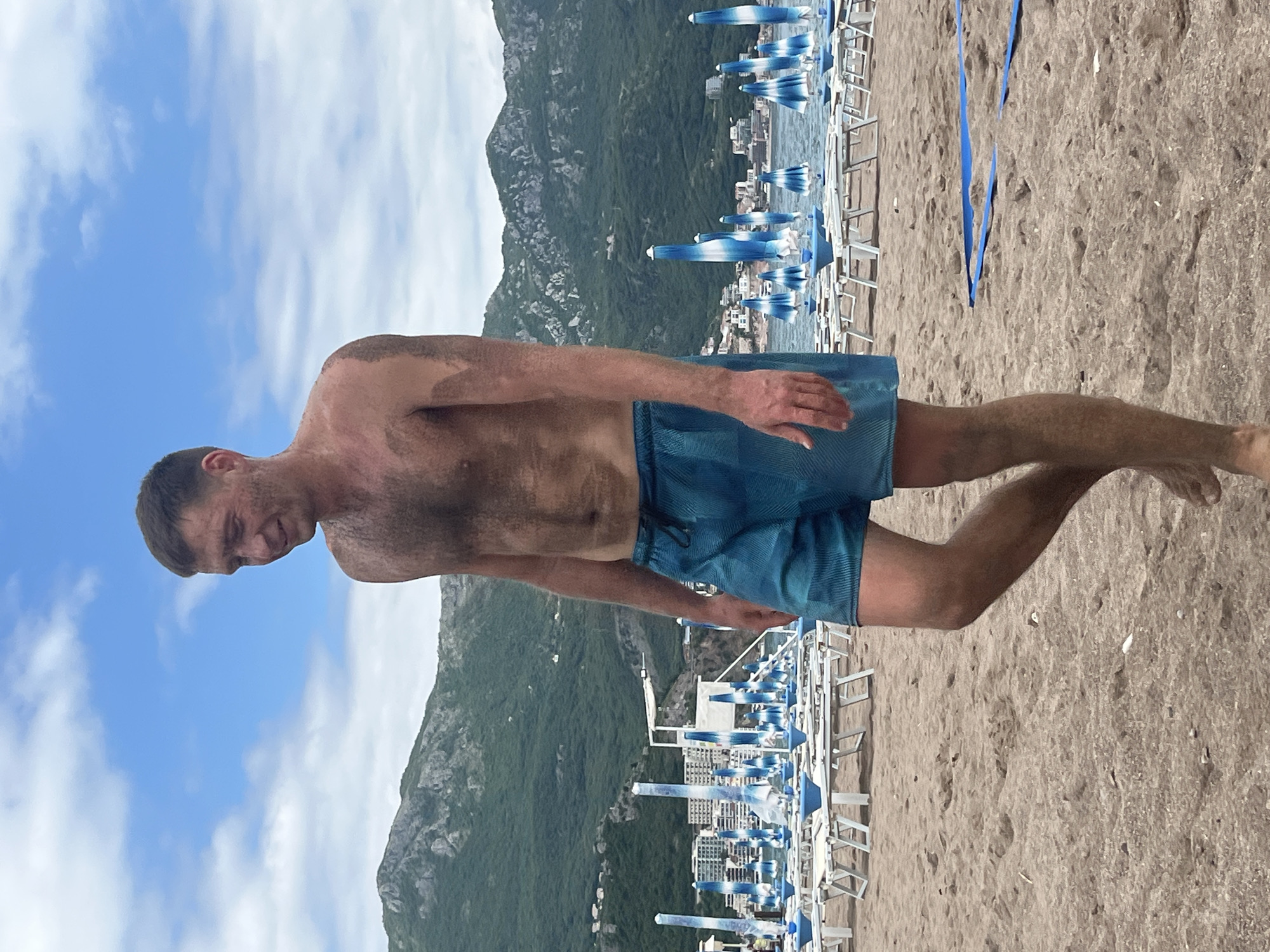 The image depicts a man standing on a sandy beach in Boreti, Montenegro. He is wearing blue swimming trunks and is shirtless. The beach is filled with rows of beach chairs and blue and white umbrellas. Behind the beach, there is a mountain covered in greenery and some buildings are visible. The sky is blue with some clouds.