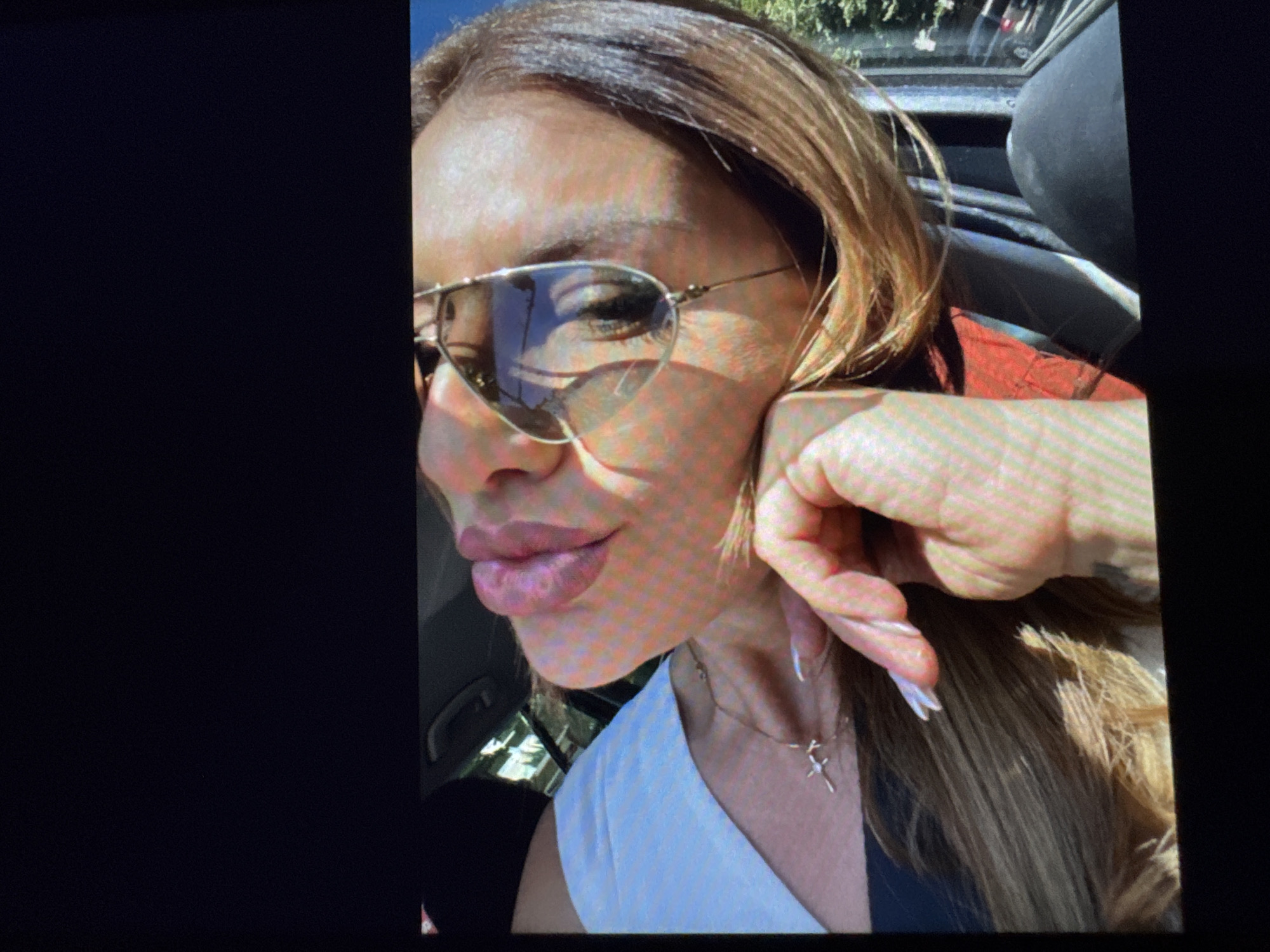 The image shows a close-up of a woman's face, tilted upwards and to the side, with her eyes closed and lips slightly parted. She is wearing aviator sunglasses and a light-colored t-shirt. A delicate silver necklace with a cross pendant is visible on her neck. Her hand is resting on her cheek, with long, manicured nails. Her hair is long and appears to be light brown. The background is blurred but suggests an outdoor setting, possibly with trees and a car visible through a window. No specific location in Plovdiv, Bulgaria can be identified from this image.