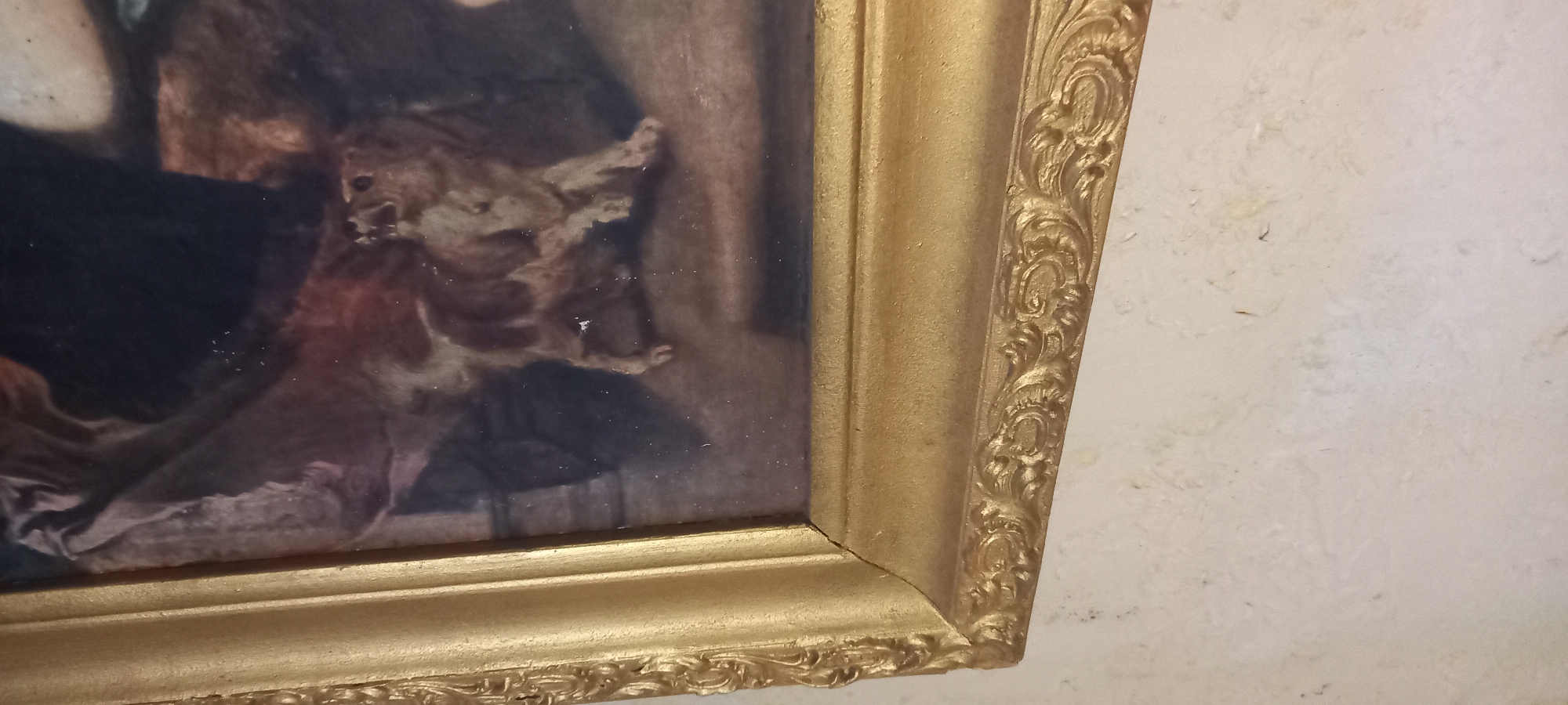 This is a close-up, eye-level, medium shot of a painting in a gilded frame. The painting depicts a small dog with brown and white markings. The dog is in a somewhat hunched or crawling posture, with its head turned to the left. The background of the painting is dark and appears to be an indoor scene, possibly with fabric or drapery visible. The frame is ornate and golden, with detailed carvings along its edge. The painting is hung on a textured, light-colored wall. The location is not discernible from the image, and it is not possible to determine if it is in Kryvyi Rih, Ukraine, based on the visual information provided.