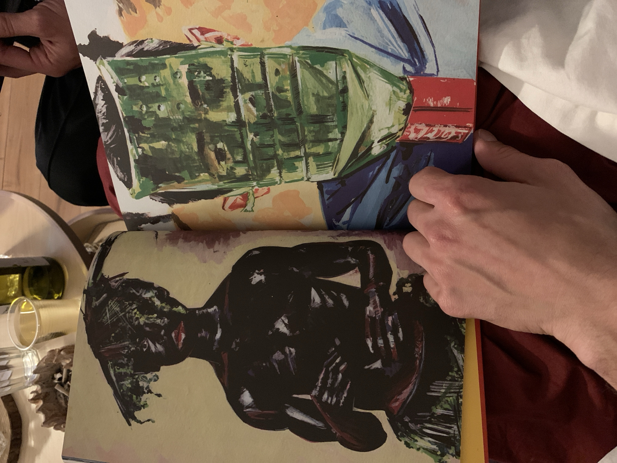 The image shows a book open with two illustrations. The top illustration is a green object with a red band, against a background of peach and blue. The bottom illustration is a dark figure lying down with arms crossed over the body, against a background of yellow. The open book is being held by a hand. Part of a white pillow and a dark red object are visible next to the hand. On a table next to the book are a wine bottle, glass and an object of organic matter. It is not possible to determine the location is in Tel-Aviv, Israel.