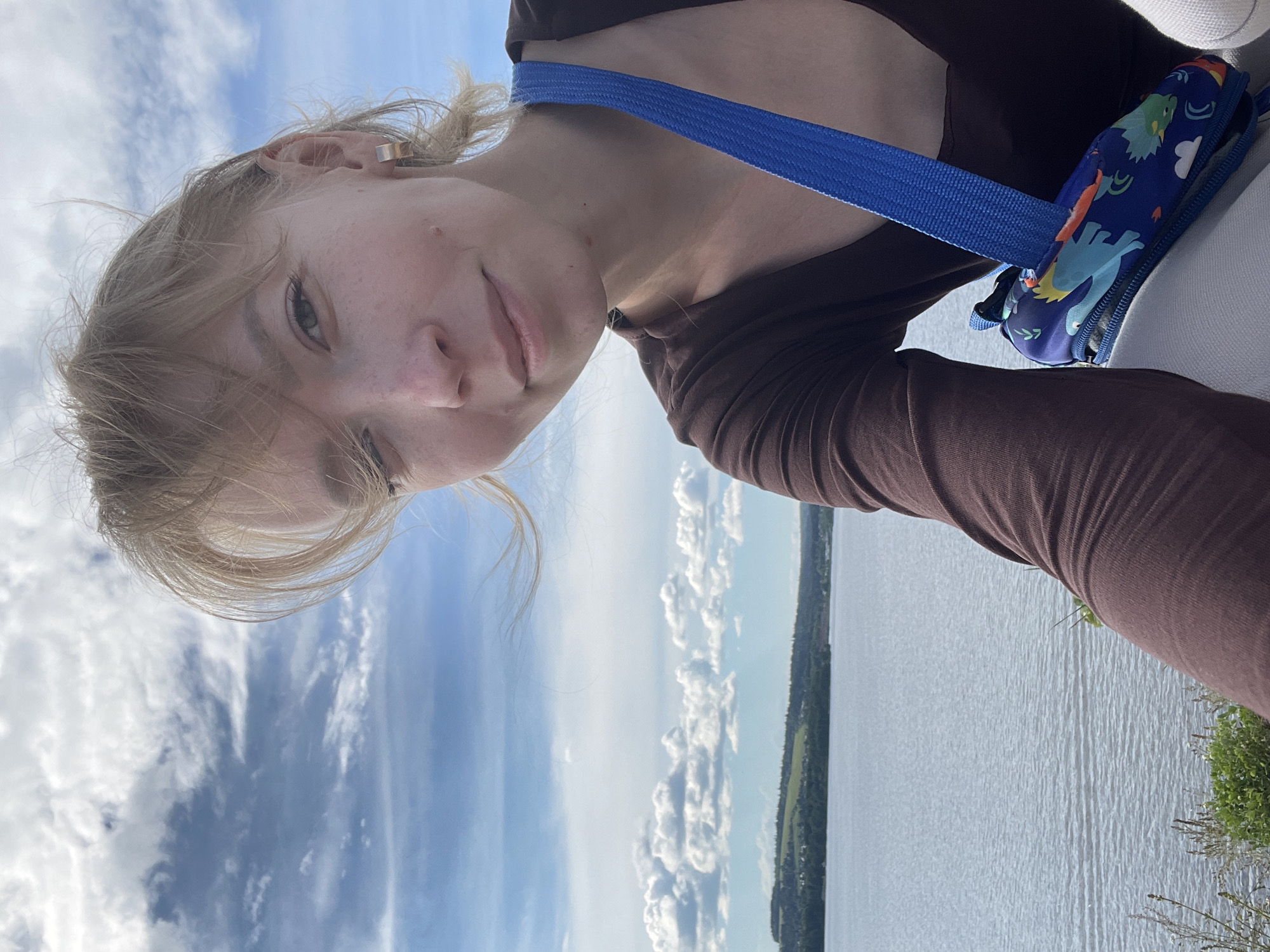 A young woman is captured in a low-angle selfie, looking upwards at the camera with a neutral to slightly smiling expression. She has blonde hair, with some strands appearing windswept, and light-colored eyes. She is wearing a form-fitting, dark brown long-sleeved top. A bright blue strap extends across her chest, to which a small blue pouch or bag is attached. This pouch features colorful, cartoon-like illustrations of dinosaurs and clouds.
The setting is outdoors under a bright, partly cloudy blue sky, with scattered white cumulus clouds. Behind the woman, a wide expanse of water, likely a large lake or river, stretches out, displaying subtle ripples on its surface. In the distance, a narrow strip of land with a dark green treeline is visible along the horizon. The lighting indicates it is daytime, suggesting clear and fair weather. The scene portrays a tranquil natural environment, located in Пятый километр, Russia. No visible text is present in the image.