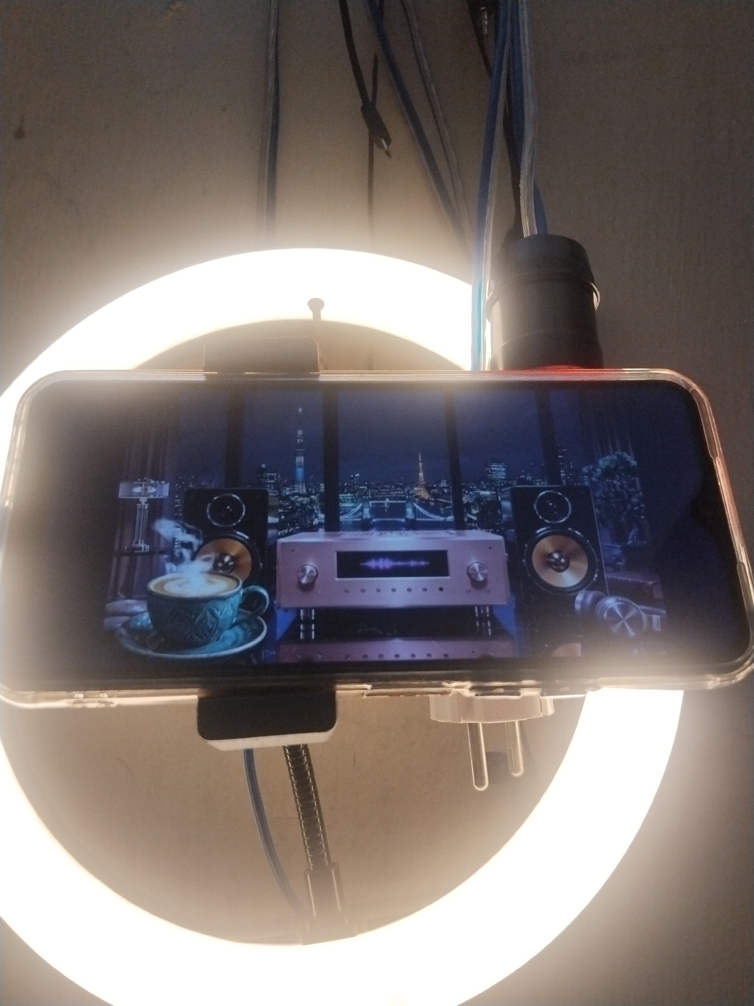 The image shows a smartphone displaying a scene with a cup of steaming coffee, two speakers, and an audio amplifier. Behind these objects, a city skyline at night is visible through a window. The smartphone is mounted on a ring light, and there are cables hanging from the ceiling. The background is a plain wall. There is no indication of the city of Tasikmalaya, Indonesia in the image.
