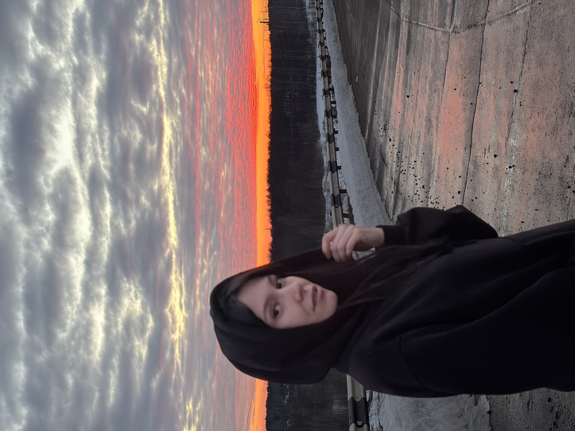 An outdoor scene in Chaikovsky, Russia, depicts a young female standing on a paved road or bridge under a dramatic sunset sky. The individual, positioned in the foreground and slightly left of center, wears a black hooded sweatshirt with the hood partially covering her dark hair. She looks towards the viewer, with her head slightly tilted and her left hand (viewer's right) raised to her ear, possibly adjusting the hood.
The road surface is grey, appearing wet in patches that reflect the intense red and orange hues of the sky, and is scattered with dark debris. Along the right side of the road, a white and dark metal crash barrier is present, behind which piles of dirty snow are visible, indicating a cold season. The sky, dominating the upper portion of the frame, transitions from a vibrant horizontal band of red and orange at the horizon to layers of grey, white, and pale yellow altocumulus clouds above. A dark silhouette of leafless trees forms the distant horizon. Faint residential structures are visible in the far left background beyond the trees, and industrial elements, possibly power lines or towers, are discernible in the far right distance. The road curves gently to the right into the mid-ground.