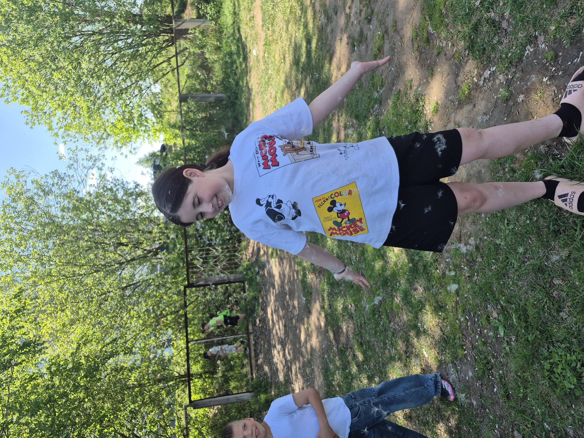 The image captures a daytime scene in what appears to be a grassy, tree-filled outdoor area, possibly in a park or a garden in the city of Chaikovsky, Russia. A young girl stands in the center, her arms extended. She is wearing a white t-shirt with cartoon graphics, black shorts, black socks, and pink Adidas-branded sandals. Fluffy white particles are scattered around her, clinging to her hair, face, and clothing. These particles appear to be dandelion seeds. In the background, partially obscured by a simple metal fence, are two figures on the ground. The lower portion of another person is visible at the bottom of the frame, wearing a white shirt and blue jeans. The ground consists of a mixture of dirt, grass, and scattered debris. Trees with green foliage form a canopy above the scene, and the sky is visible through the branches.