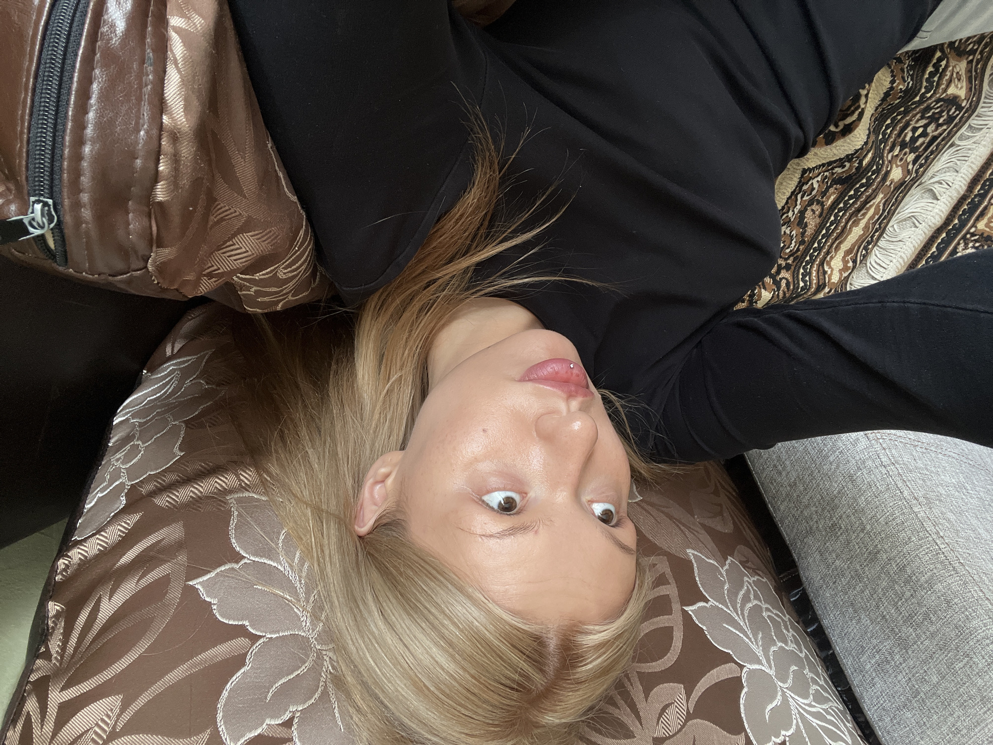 A woman with blonde hair is lying upside down on a couch. She is wearing a black long-sleeved shirt. The couch has multiple patterned cushions: one with brown floral designs, another with brown and beige stripes, and a third in plain gray. The image appears to have been taken indoors in Chaikovsky, Russia.