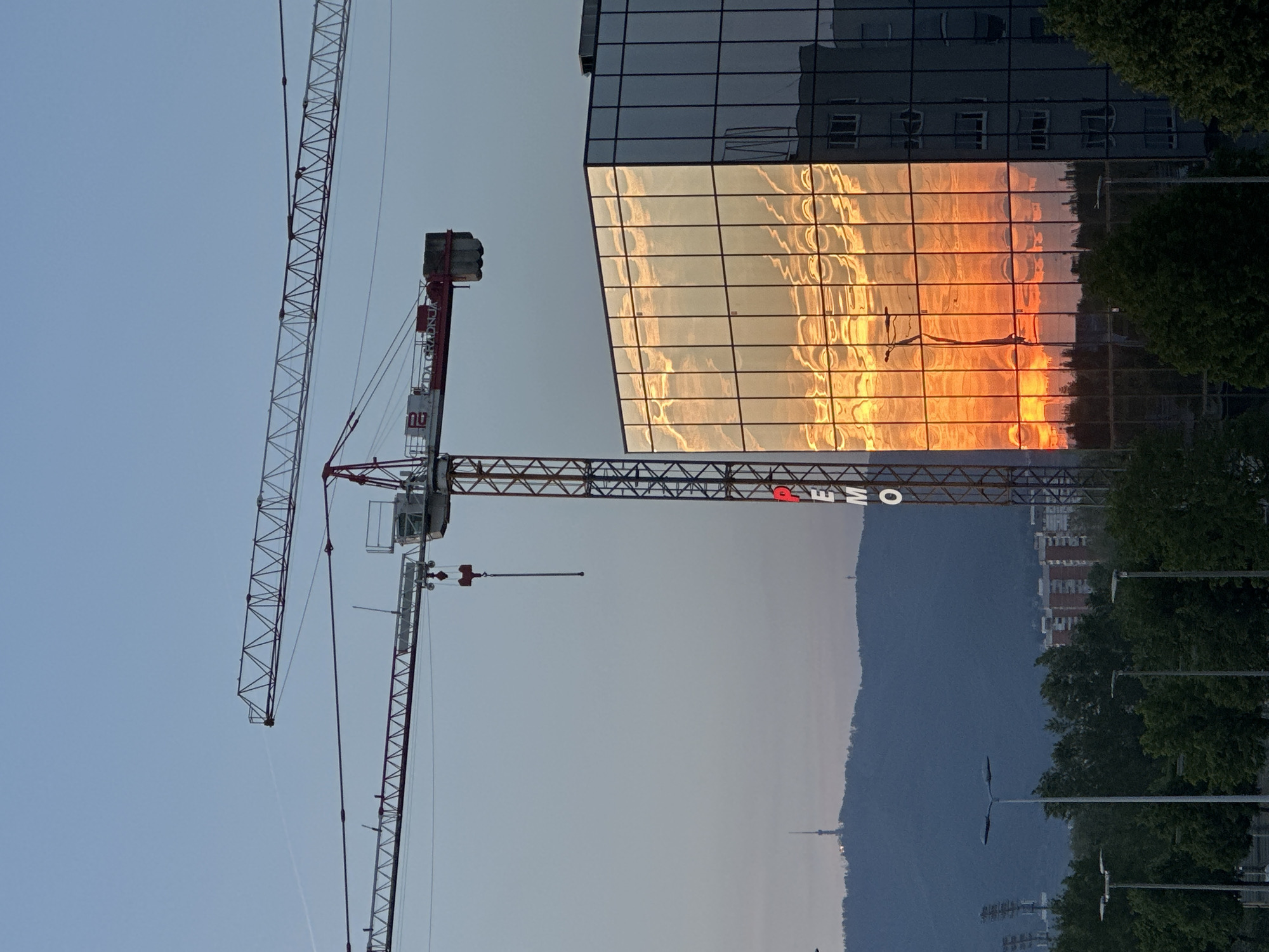 The image depicts a cityscape in Zagreb, Croatia at dusk or dawn. A modern office building with a glass facade reflects an orange sunset. A construction crane stands in the foreground against a gray sky. In the background, a dark, sloping hill is visible, with a television tower atop it. Trees line the lower right of the frame, and streetlights are positioned adjacent to the trees.