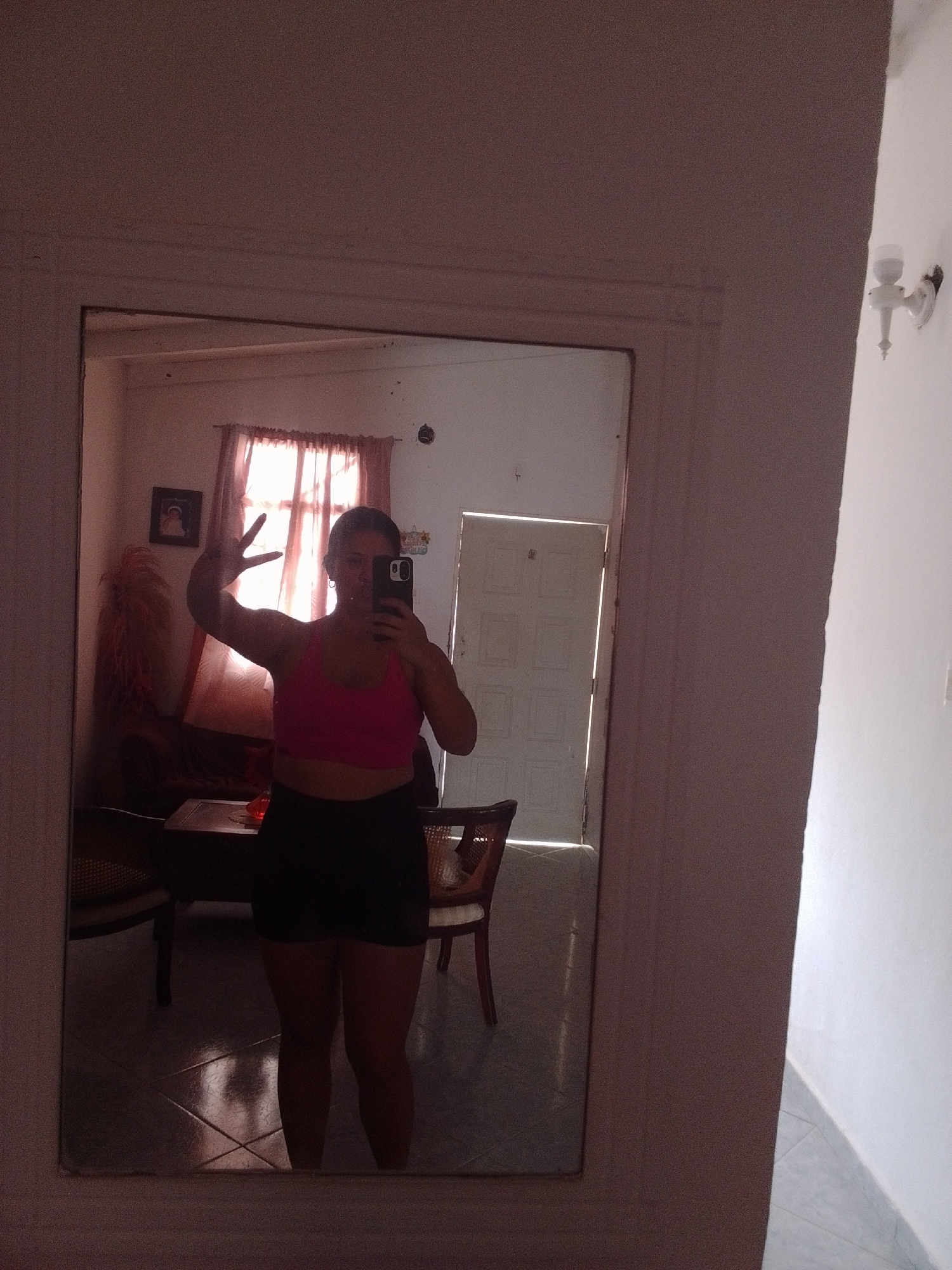 The media file shows a young woman taking a mirror selfie in what appears to be a residential interior. She is wearing a pink sports bra and black shorts. She is holding her phone in her right hand and has her left hand raised, fingers spread, in a peace sign. Behind her, the reflection in the mirror reveals a room with a window covered by sheer pink curtains, a dark wooden table, and chairs made of woven material. A small framed picture is visible on the wall near the window, and what looks like a decorative fluffy object is also present. The floor is tiled and reflects the light coming from the window. A doorway with light spilling through it is visible in the background reflection. The lighting suggests it might be daytime, possibly late afternoon due to the warm light. The overall impression is casual and domestic.