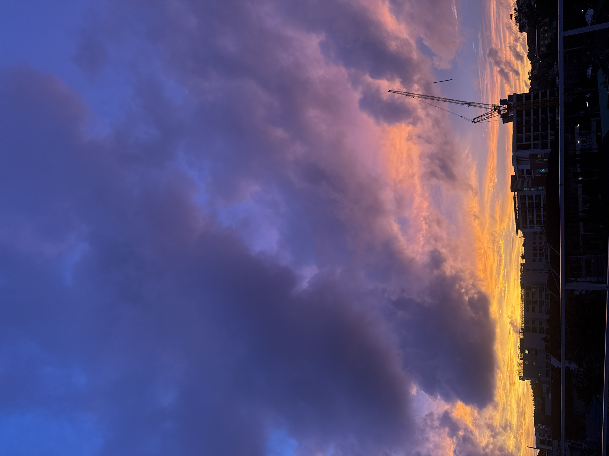 A dramatic sunset in Karon, Thailand, paints the sky with vibrant hues of orange, pink, and deep blue. Dark, heavy clouds dominate the upper portion of the frame, contrasting with the fiery glow at the horizon. Silhouetted against this colorful sky are the dark outlines of buildings and a construction crane, suggesting ongoing development in the area. The foreground features a balcony railing, implying the image was taken from a viewpoint overlooking the cityscape. The overall scene captures the beauty of a tropical sunset juxtaposed with urban structures.