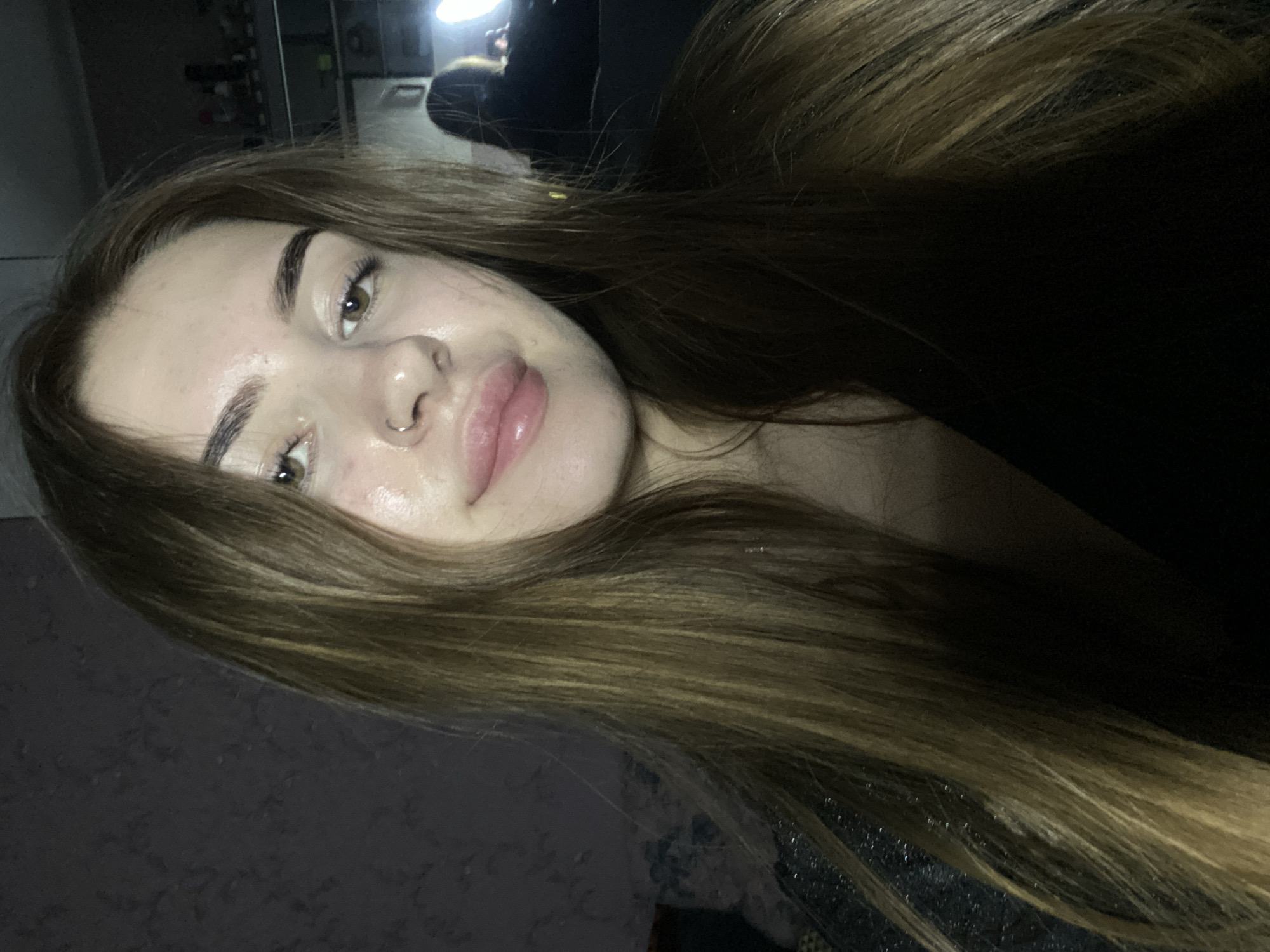 The image features a young woman captured in a medium close-up, oriented diagonally across the frame. She has long, straight, medium-brown hair with lighter highlights that frames her face and cascades over her shoulders. Her complexion appears dewy, and she has light brown eyes, dark, well-defined eyebrows, and full, slightly parted lips. A small hoop piercing is visible on her left nostril. She wears a dark top, with a subtle glittery texture visible on her right shoulder/chest area. Her expression is neutral, with a slight hint of a smile as she looks directly at the camera, suggesting she is posing for a selfie.
The background is dark and indistinct, indicating an indoor setting, possibly dimly lit. A bright, circular artificial light source is visible in the upper background, creating highlights on her hair and face and casting some shadows. No text is visible in the image.