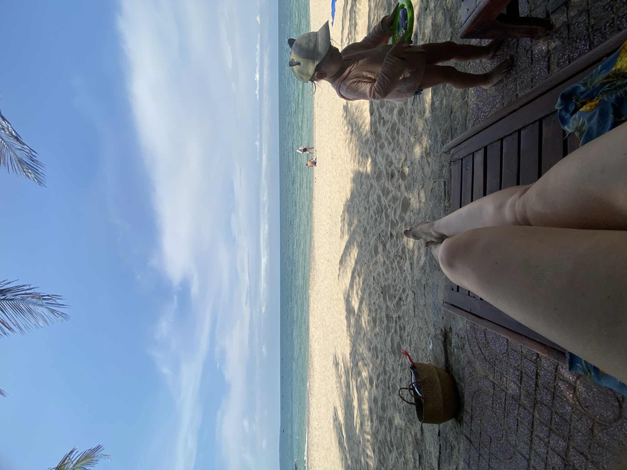 A woman's legs are visible in the foreground, resting on a wooden lounge chair on a tiled pathway. To the left, a woven basket sits on the sand, containing a bottle and a red object. A child wearing a cap and a light pink shirt stands on the sand to the right, holding a green frisbee. In the background, the calm sea meets a sandy beach with a few distant figures. Palm fronds cast shadows on the sand, indicating a tropical or subtropical location. The sky is blue with scattered clouds. The scene appears to be a resort or beach area in Phu Thuy Ward, Vietnam.