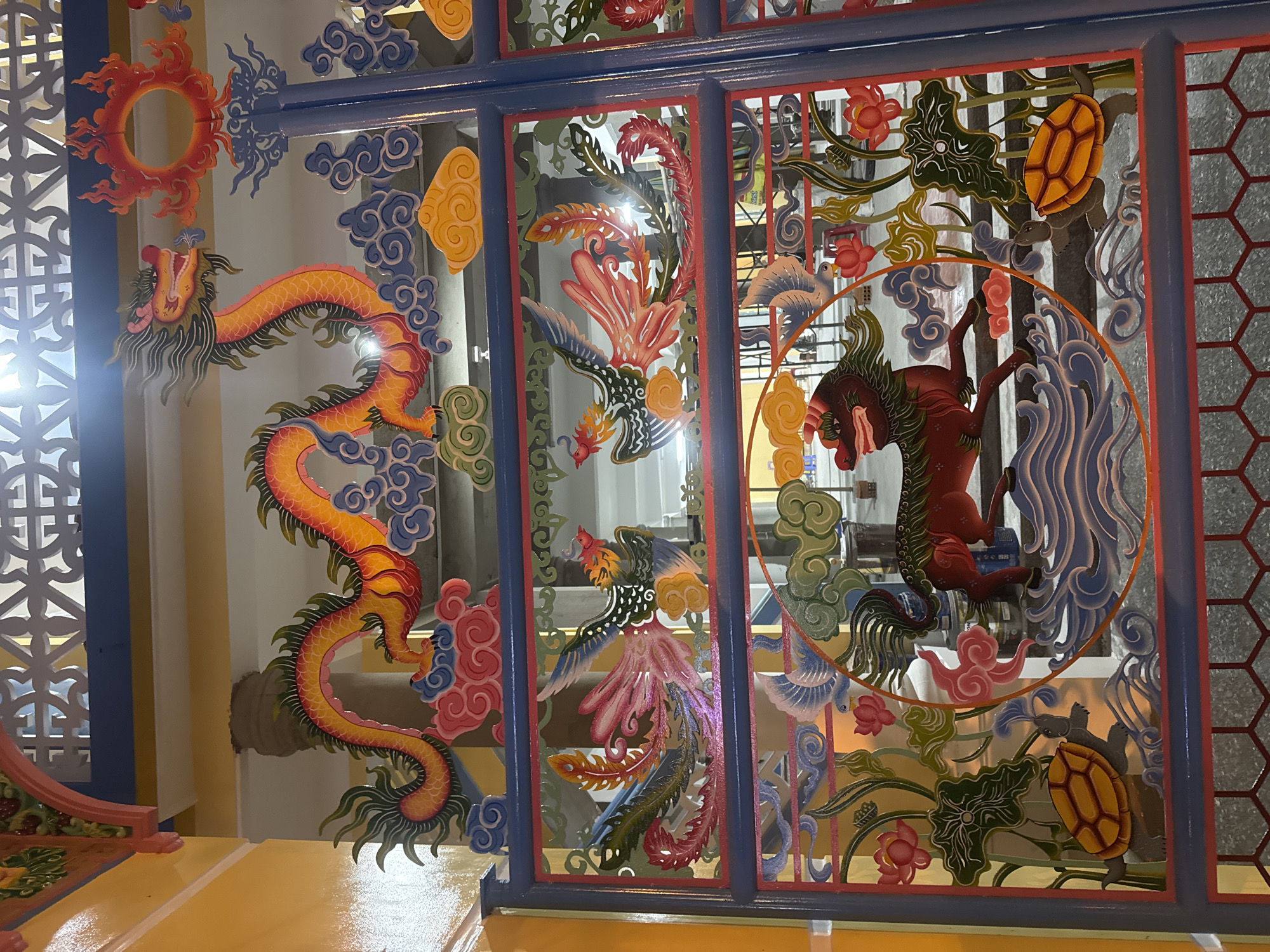 The image shows a painted screen with several panels. The panels depict stylized creatures: a dragon, a phoenix, a Qilin (a mythical hooved Chinese chimerical creature) and a turtle. The screen has a blue frame, with red accents around the panels. The screen is positioned on a yellow surface. The image appears to be taken indoors, but the exact location in Nha Trang Ward, Vietnam is not identifiable from the image.