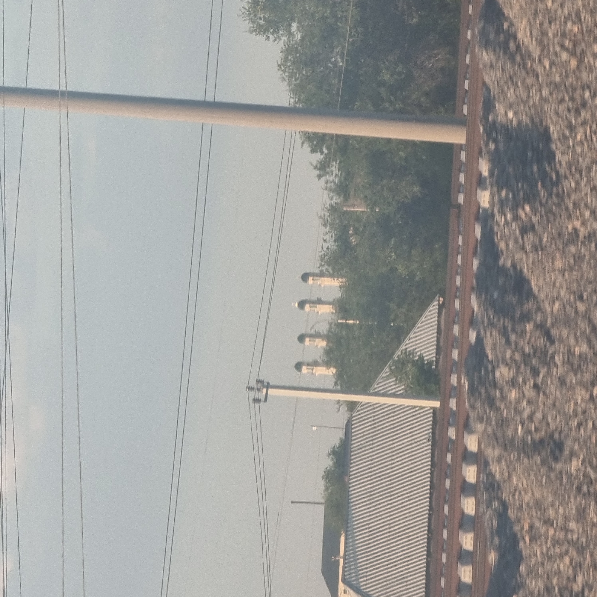 The image shows a view from a moving vehicle on a clear day in the city of Arys, Kazakhstan. In the foreground, a railway track with ballast and concrete sleepers is visible. Behind the railway, a building with a corrugated metal roof and a black roof is present. Several power lines and a utility pole are also visible. In the middle ground, trees form a dense canopy. In the background, a row of minarets, characteristic of a mosque, can be seen rising above the trees. The sky is overcast with a pale blue hue, and sunlight casts shadows on the ground. The image appears to have been taken during daytime, likely in the afternoon, given the lighting and the presence of shadows. There are no people or animals visible in the frame.