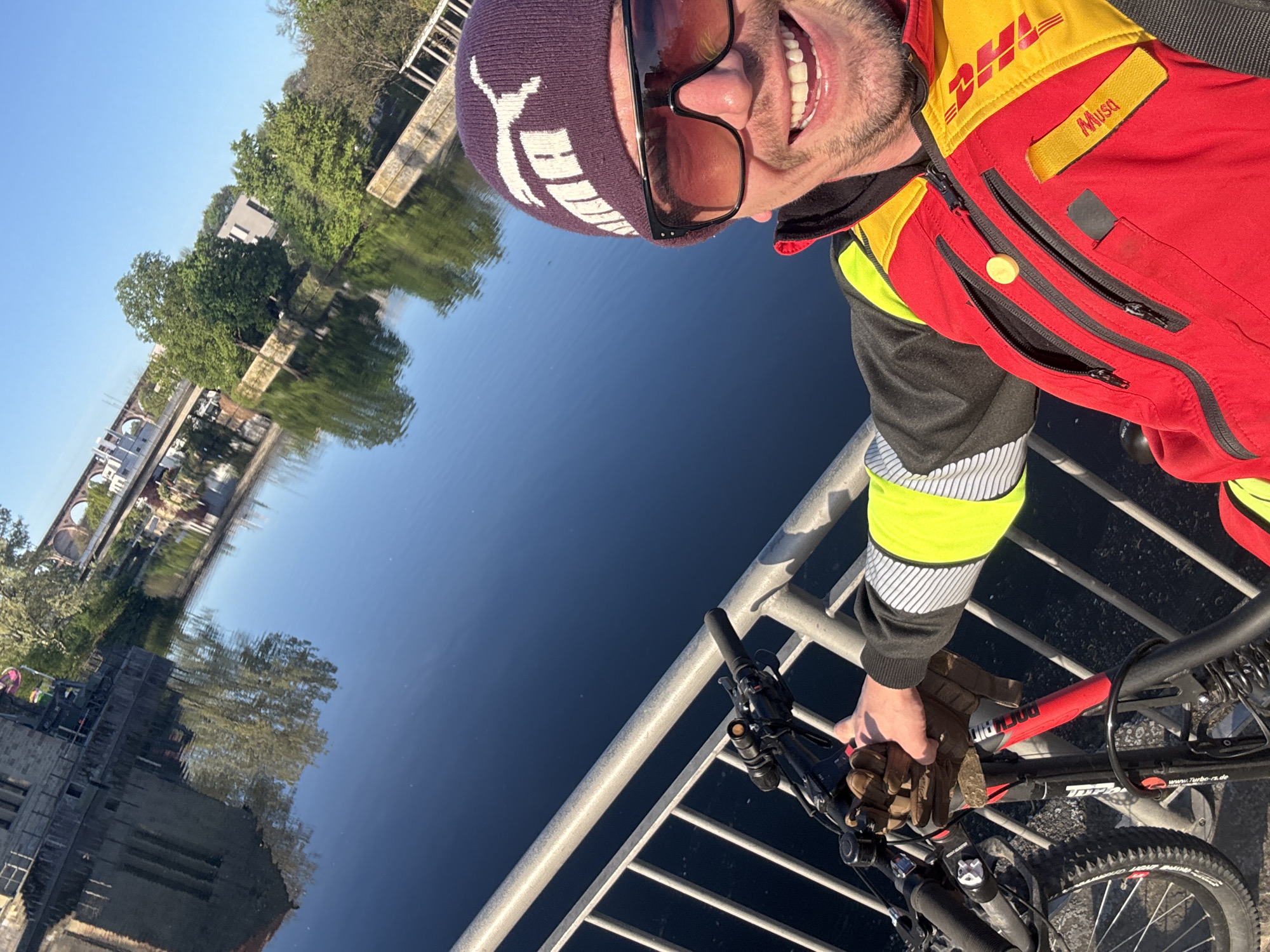A man wearing a DHL uniform, a purple Puma beanie, and sunglasses is on a bridge with his bicycle. He is smiling into the camera. He is standing on the bridge that runs over the Enz River in Bietigheim-Bissingen, Germany. The reflection of buildings, trees, and an arched viaduct is visible in the water. The man is wearing brown gloves and holding the handlebars of his red and black mountain bike.