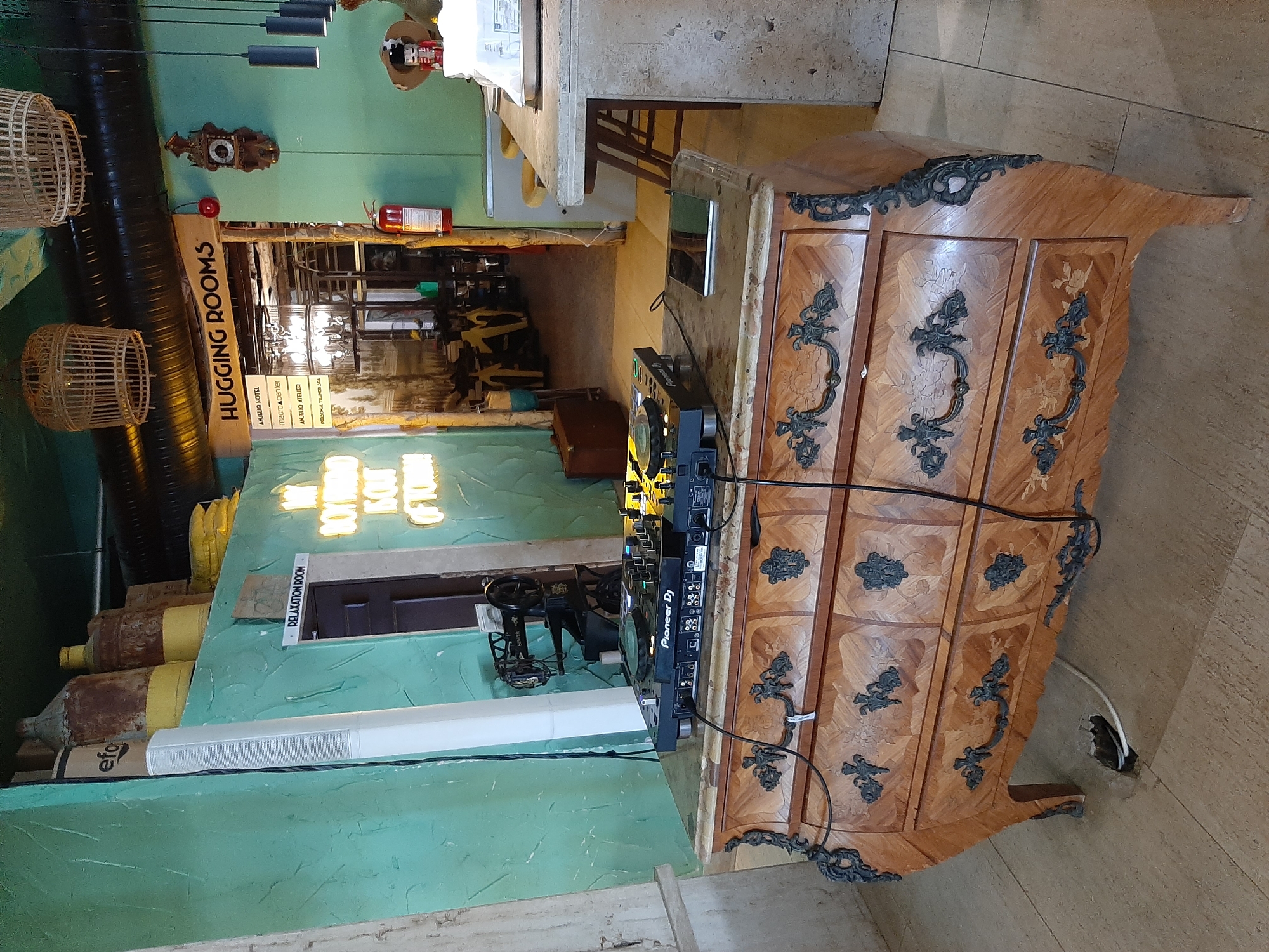 The image shows a DJ setup on top of an ornate wooden dresser in what appears to be a commercial establishment. The DJ controller, a Pioneer DJ model, is connected to various cables. A tablet computer is also placed on the dresser. In the background, there is a wall painted a teal color with various decorative elements, including a neon sign that reads "MY BODY IS OUT OF TOWN." Further in the background, an archway leads to another section of the establishment, where a sign reads "HUGGING ROOMS." Other objects visible include industrial-looking pipes, hanging light fixtures, a fire extinguisher, and a vintage sewing machine. The floor is tiled. There are no people clearly visible in the scene. The establishment seems to be located in Alanya, Turkey, as indicated by the text on a sign visible in the background which lists various businesses including "AMELIO HOTEL".