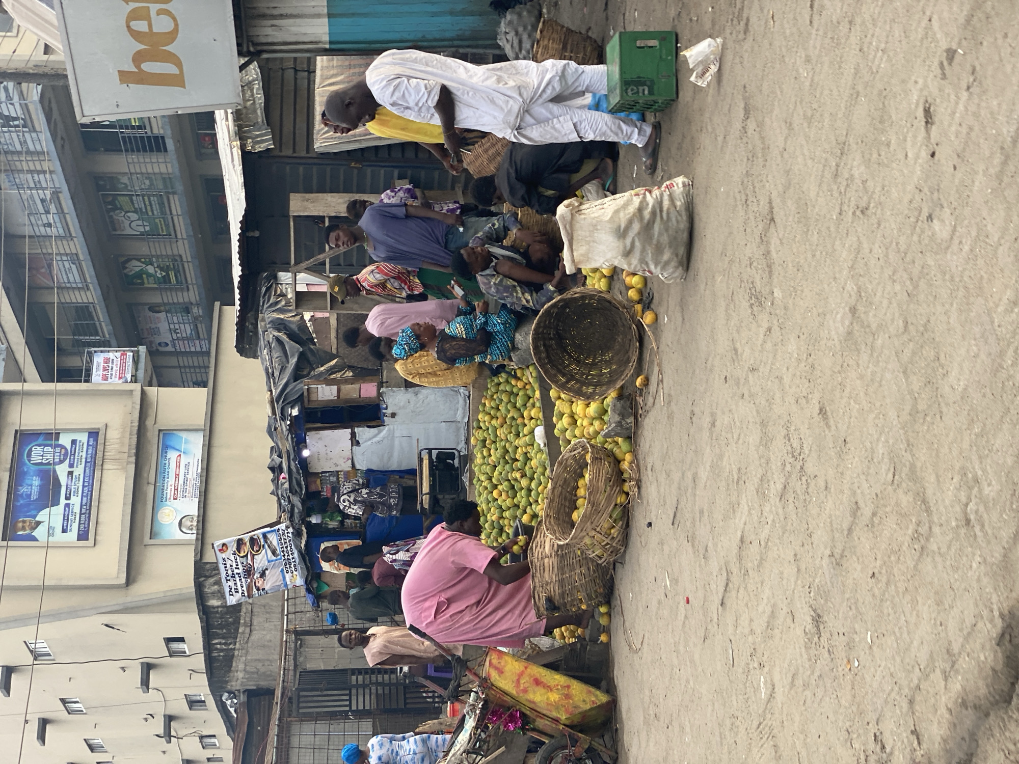 This image captures a vibrant street market scene in an urban area, likely Ado, Nigeria. The setting features a bustling street with a dirt or concrete ground, flanked by multi-story buildings on the left and makeshift stalls on the right.
Numerous people, appearing to be vendors and customers, populate the scene. Many individuals are seated or squatting on the ground, attending to their wares. A man in a white traditional attire stands prominently on the left, next to a green crate. Another person in a pink top is seen kneeling beside a large pile of yellow-green fruits, suggesting they are a fruit vendor. Other people are standing or moving between the stalls, engaged in various market activities.
The primary activity depicted is commercial exchange within an outdoor market. Vendors are selling large quantities of fruits, predominantly yellow-green mangoes or oranges, displayed in large piles on the ground and in numerous woven baskets. Other objects include a white sack, a yellow and red wheelbarrow, and the green "Heineken" (partially visible as "Ken") crate. The makeshift stalls are constructed from tarpaulins and corrugated metal sheets, typical of informal markets.
Notable details include the daytime setting, indicated by bright, diffused light, suggesting an overcast day or an early/late time. Several signs are visible, providing contextual clues. On a building, a partial sign reads "be," and another says "WOR SHIP WITH US" with an address "2ND FLOOR, VIEW POINT PLAZA, BY KYZ COMPLEX, NEPA ROAD, AJAR." A sign for "FOUNDATION FAITH CHURCH" is also seen. A banner announces "De Toniz Barber/ Dread luck shop" with phone numbers (07034419555, 070710437, 0911400573). A small sign also reads "HOPE LIVES HERE." The overall atmosphere appears busy and functional, characteristic of a lively market.