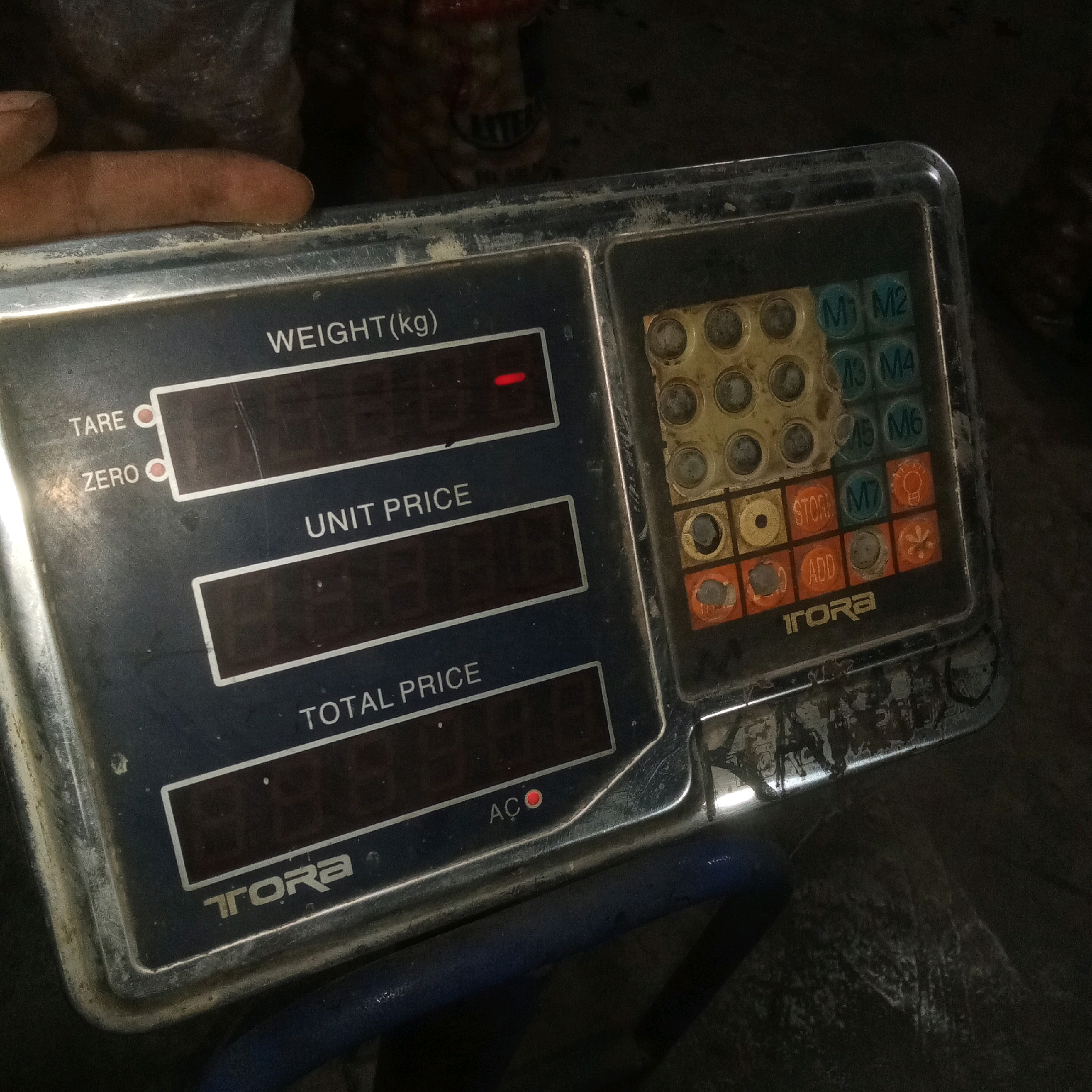 The image shows a close-up of an old digital scale, possibly in a market setting, with a hand visible on the top left edge. The scale has a dark blue face with several digital displays and a keypad with circular buttons. The labels "WEIGHT(kg)", "UNIT PRICE", and "TOTAL PRICE" are visible above their respective displays, which are currently dark. The keypad has buttons labeled with numbers (M1 to M6) and functions such as "STORE", "ADD", and "AC". The scale has the brand name "TORA" at the bottom. The surrounding environment is dimly lit and appears somewhat dirty or dusty, with out-of-focus dark shapes in the background that could be produce or other goods. The presence of this type of scale suggests a commercial transaction, likely a small business or market stall. Given the context of Karawang, Indonesia, this scene is likely located in a traditional market, roadside stall, or a small shop within the city.