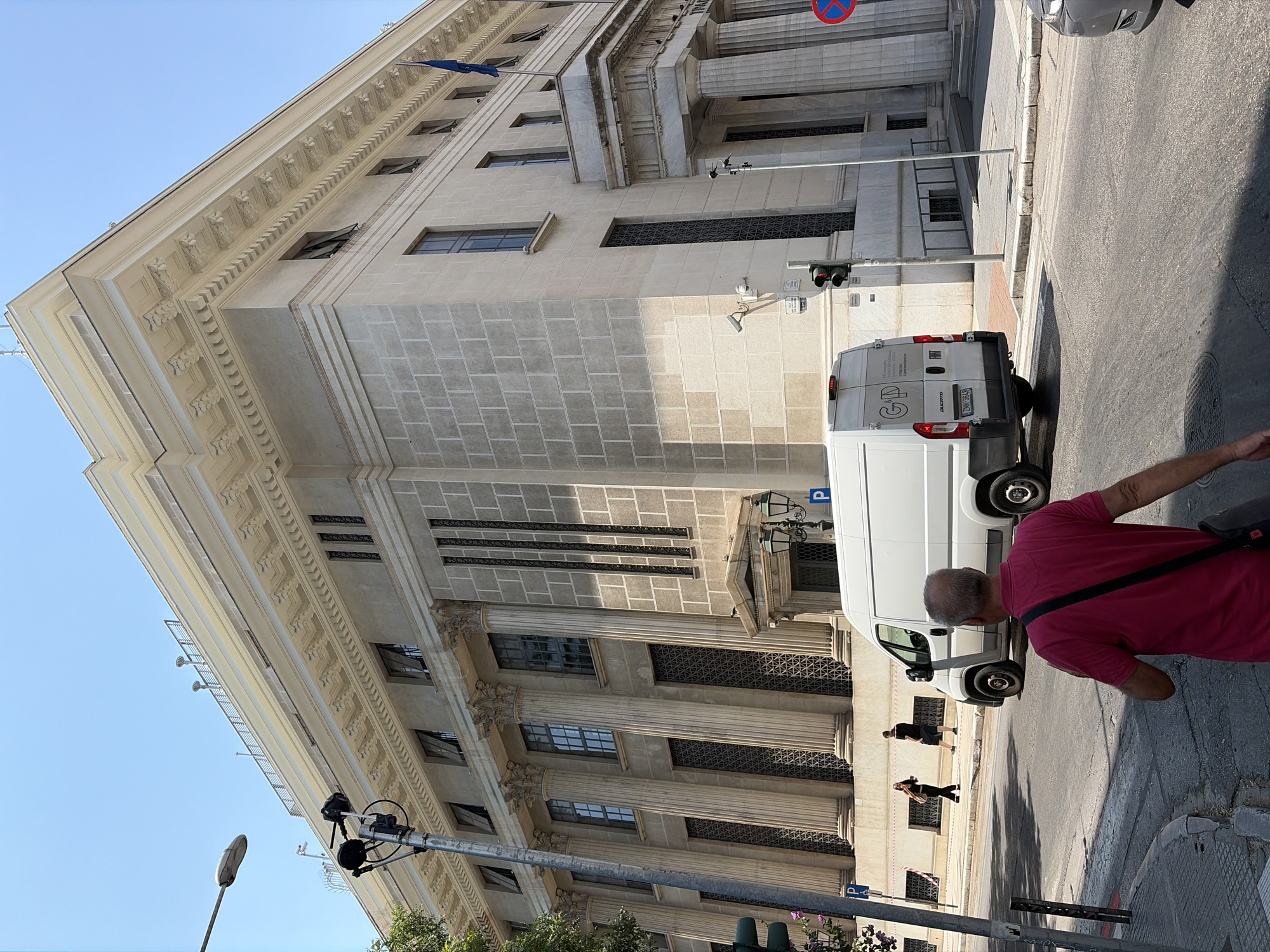 The image shows the exterior of the Bank of Greece building in Thessaloniki, Greece, on a sunny day. The building is a large, neoclassical structure with a light-colored stone facade. It features imposing columns, arched windows with ornate grills, and a decorative cornice. A blue flag flies from a flagpole on the roof.
In the foreground, a white van is parked on the street in front of the building, with its rear facing the camera. A man wearing a pink polo shirt and a black bag slung across his shoulder is walking away from the camera on the sidewalk, obscuring the lower portion of the van. Across the street, two individuals are walking. One is a woman in dark clothing, and the other is a man in a dark t-shirt and shorts.
Street furniture visible includes a traffic light, a street lamp, and parking signs. The street is paved with asphalt, and there are sidewalks on either side. Trees are visible on the left side of the image. The sky is clear and blue. The overall impression is a street scene in a European city, dominated by the grand architecture of the bank.