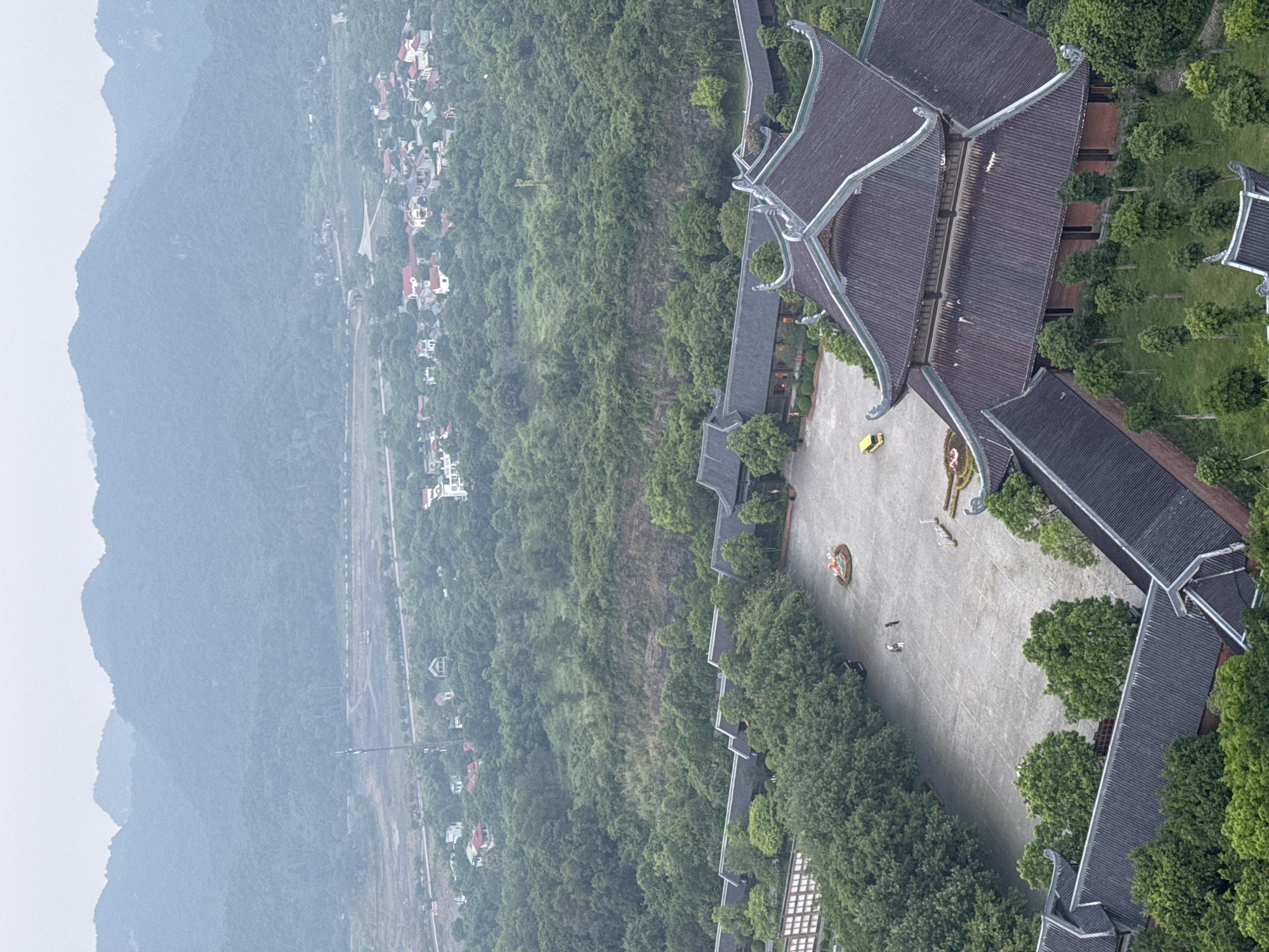 This aerial photograph depicts a traditional Vietnamese temple complex nestled amidst lush green hills and a distant village. The temple architecture features dark, tiled roofs with ornate, upturned eaves, set against a backdrop of rolling mountains shrouded in a hazy, overcast sky. In the foreground, the temple courtyards are paved with stone, and a few scattered trees provide shade. There are two figures walking in the courtyard, and a yellow vehicle parked nearby. Beyond the temple, a small village with red-tiled roofs is visible, surrounded by dense vegetation. The overall scene conveys a sense of tranquility and cultural heritage.