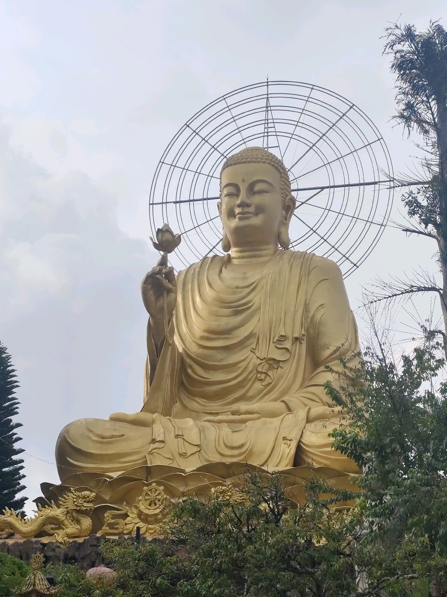 The image shows a large, golden statue of the Buddha in a seated position, holding a lotus flower in his right hand. The statue is adorned with a halo behind its head. At the base of the statue, there are decorative elements including a dragon sculpture and a dharma wheel. The statue is surrounded by lush green trees. The sky above is cloudy. This is likely the Thien Vien Truc Lam monastery in Đà Lạt, Vietnam, a prominent spiritual site in the Lâm Viên plateau region.