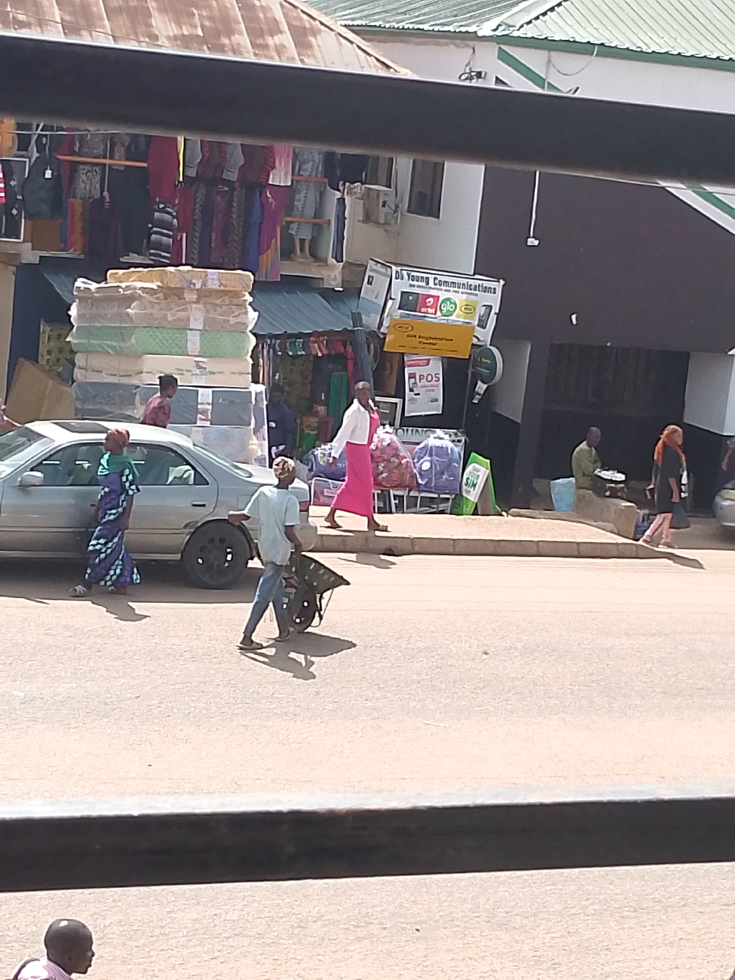 This is a street scene in Bukuru, Nigeria, likely during the daytime. A silver sedan is parked at the side of a paved road. To the right of the car, a woman in a long pink dress and white jacket walks away from the camera. Further down the sidewalk, a man in a green shirt sits on a low wall. To the left of the car, a woman in a blue and white patterned dress walks towards the camera, with a person behind her on the car's roof. A man in a light blue shirt pushes a wheelbarrow across the road.
In the background, there are several shops. One shop has a display of mattresses stacked high. Another shop has clothing hanging on racks. A sign reads "Di Young Communications" with logos for mobile network operators like Airtel, Glo, and 9mobile. Another sign indicates "SIM Registration Center." A green sign with "SIM" is also visible. The overall atmosphere appears to be that of a busy marketplace or commercial street. The weather seems sunny, casting shadows on the ground.