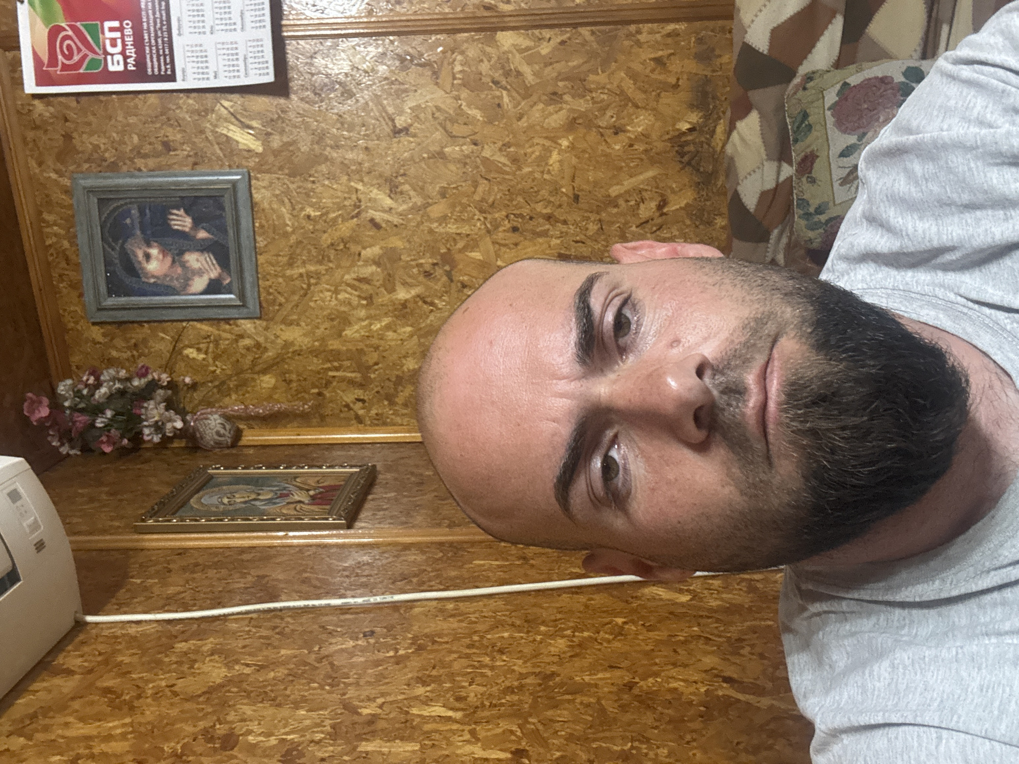 The image shows a man with a shaved head and a beard, wearing a gray t-shirt. He appears to be indoors. The background features wood-paneled walls, two framed religious icons, a small floral arrangement, and what looks like a political poster. A white cord is visible near the lower left corner, likely connected to an appliance or device. The poster has the text "БСП Раднево" which refers to the Bulgarian Socialist Party in the town of Radnevo, not Sarnevo.