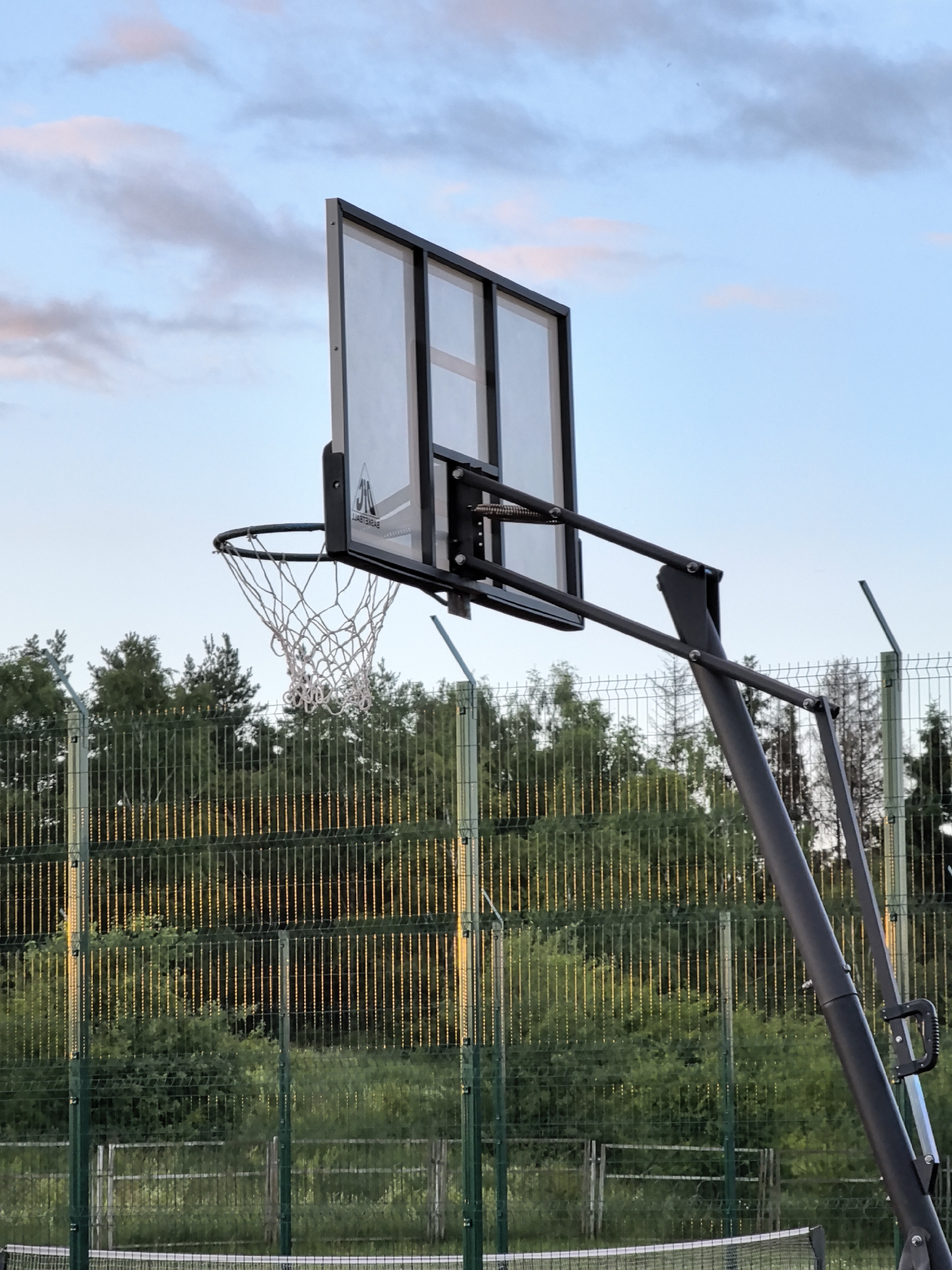 An outdoor basketball hoop with a clear backboard and white net stands against a pale blue sky with scattered pink and white clouds. The hoop is mounted on a dark metal pole. Behind the hoop, a tall green metal fence encloses a grassy area with lush green trees in the background. The setting appears to be a sports court or park.