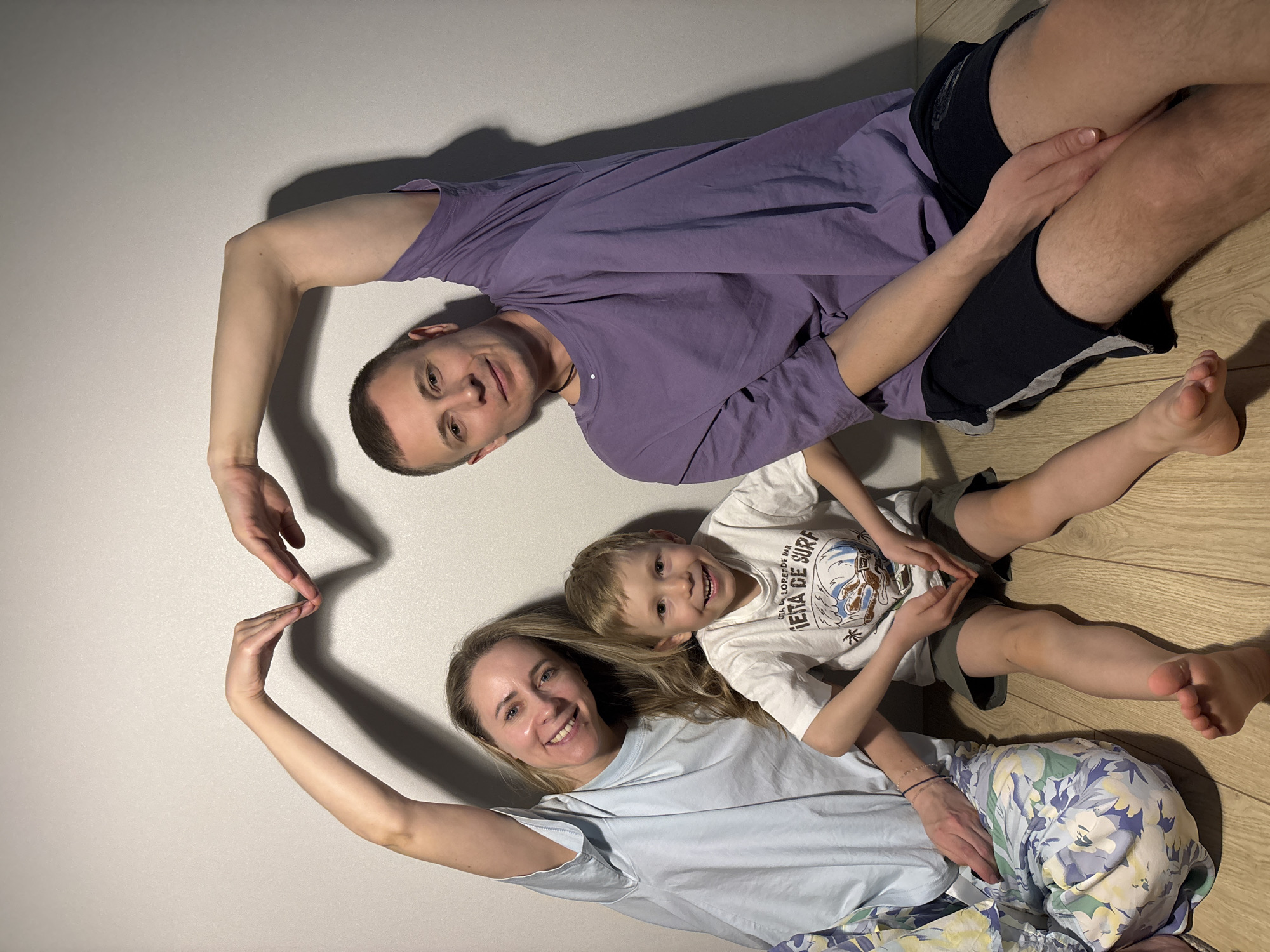 A happy family of three—an adult male, an adult female, and a young boy—are lying down and posing for a photo indoors in Chaikovsky, Russia. The adult male, wearing a purple t-shirt and dark shorts, is positioned at the top of the frame. Below him, the adult female, in a light blue t-shirt and floral-patterned pants, lies with the young boy in a white graphic t-shirt nestled beside her. The man and woman are forming a heart shape with their hands on the left side of the image, symbolizing love and unity. All three subjects are smiling brightly at the camera, conveying a sense of joy and affection. They are lying on a plain, light-colored surface, with a section of wooden floor visible on the right side of the image. Shadows cast to the right suggest an artificial light source from the left. The child's white t-shirt has visible text, including "FLORENCE NH" and "ERA DE SURF."