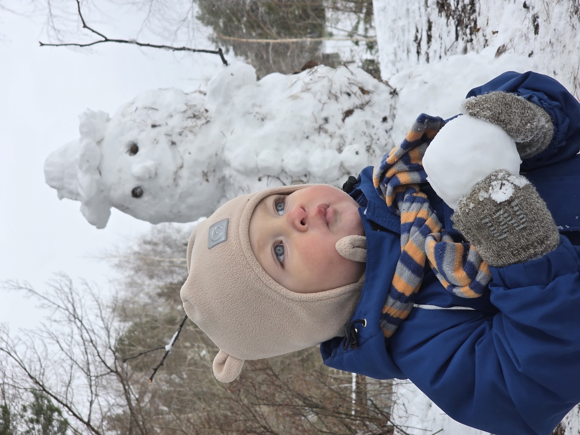 A young child, wearing a beige bear-eared hat, a blue jacket, a striped scarf, and grey mittens, is holding a snowball. The child is outdoors in a snowy environment near a snowman. The location is Chaikovsky, Russia. Bare trees are visible in the background.