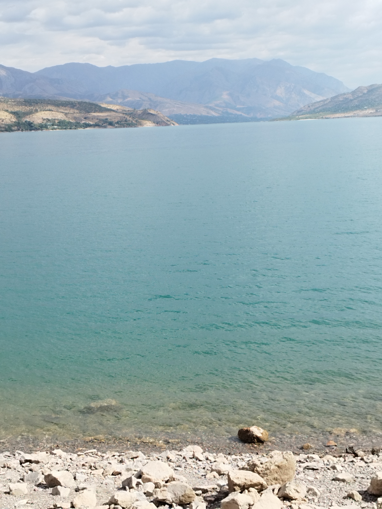 A wide, calm lake occupies the majority of the frame, its surface a shimmering turquoise. In the distance, rolling hills and mountains, layered in shades of blue and gray, recede towards the horizon under a hazy, overcast sky. The shoreline in the foreground is rocky and barren, covered in a scattering of gray pebbles and larger, pale stones. Some of these stones are partially submerged in the shallow, clear water, revealing the rocky lakebed beneath. The overall scene depicts a natural landscape, likely a reservoir or a large natural body of water surrounded by mountainous terrain. There are no people or man-made structures immediately visible in the frame.