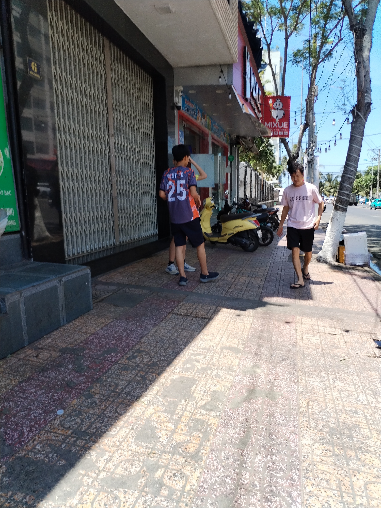 The image captures a street scene in Bac Nha Trang Ward, Vietnam. Two young people stand on the sidewalk, near a building with a security gate. One is wearing a soccer jersey with the number 25. Motorbikes are parked nearby. A third person walks on the sidewalk toward the street, wearing a t-shirt. The sidewalk is paved with patterned stones. In the background, a Mixue store sign is visible, along with trees and utility poles. The sky is clear.
