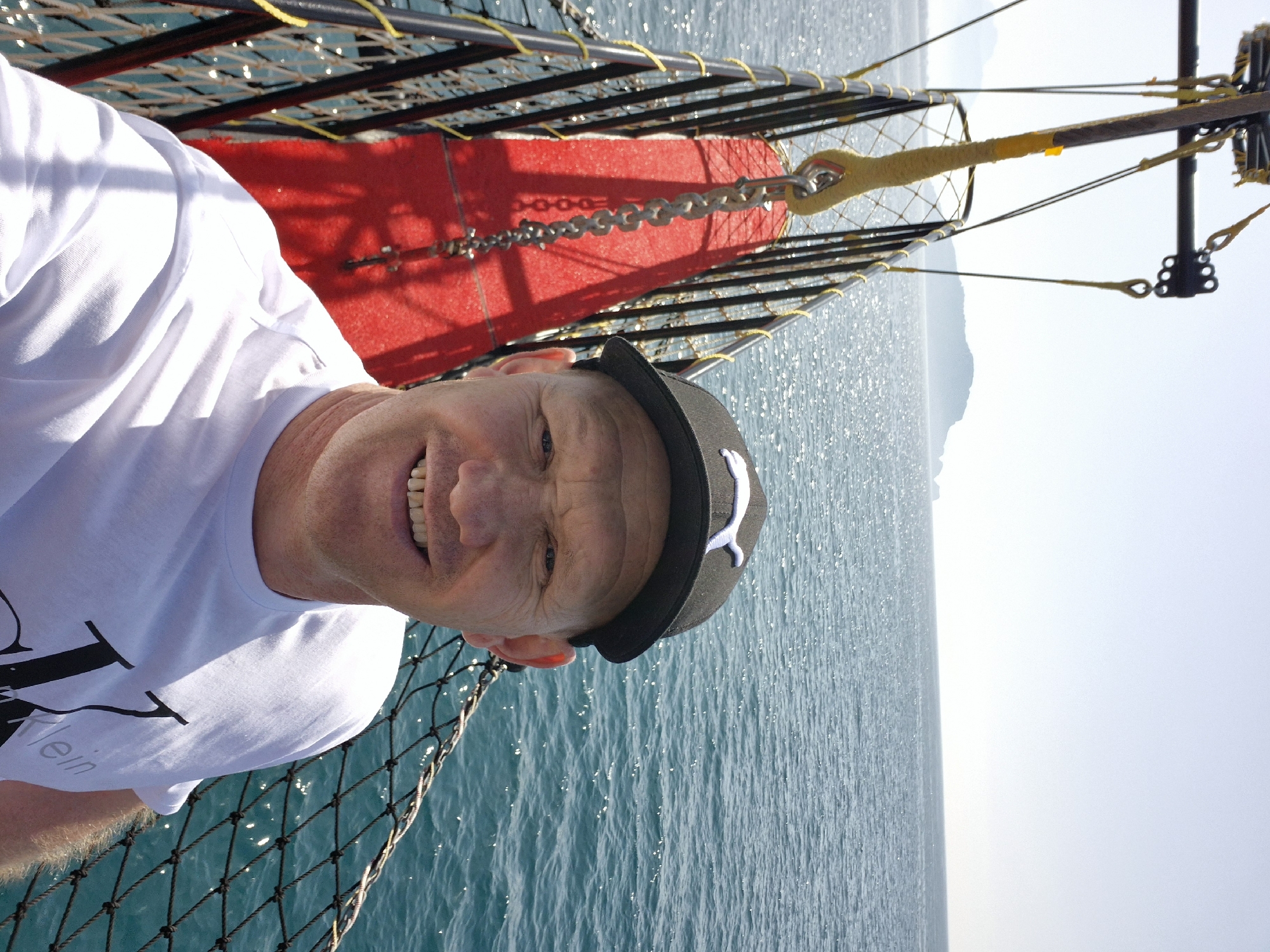 A Caucasian man wearing a black cap with a white Puma logo and a white T-shirt is on a boat, with the ocean visible in the background. The boat has a metal net and a red object attached by a chain. A coastline or mountains are visible in the far distance. The location is likely Alanya, Turkey, given the Mediterranean-like setting of the sea and distant land.