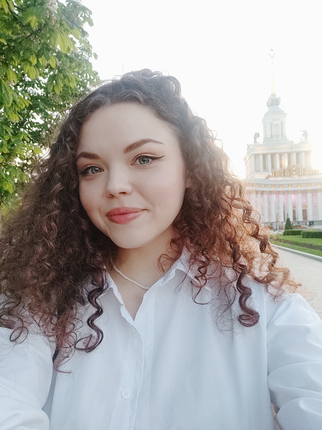 A woman with curly brown hair is the primary subject of a photograph taken outdoors. She is wearing a white, collared button-down shirt and a delicate pearl necklace. Her makeup is noticeable, featuring defined eyeliner and lip color. In the background, a building is visible. It is characterized by a tall, ornate central tower with a spire, typical of Soviet-era architecture. The building is the Central Pavilion of VDNKh, located in Moscow, Russia. Green trees are present in the upper left corner of the frame. The background suggests a park or garden setting. The lighting is soft, implying it may be late afternoon or early evening.