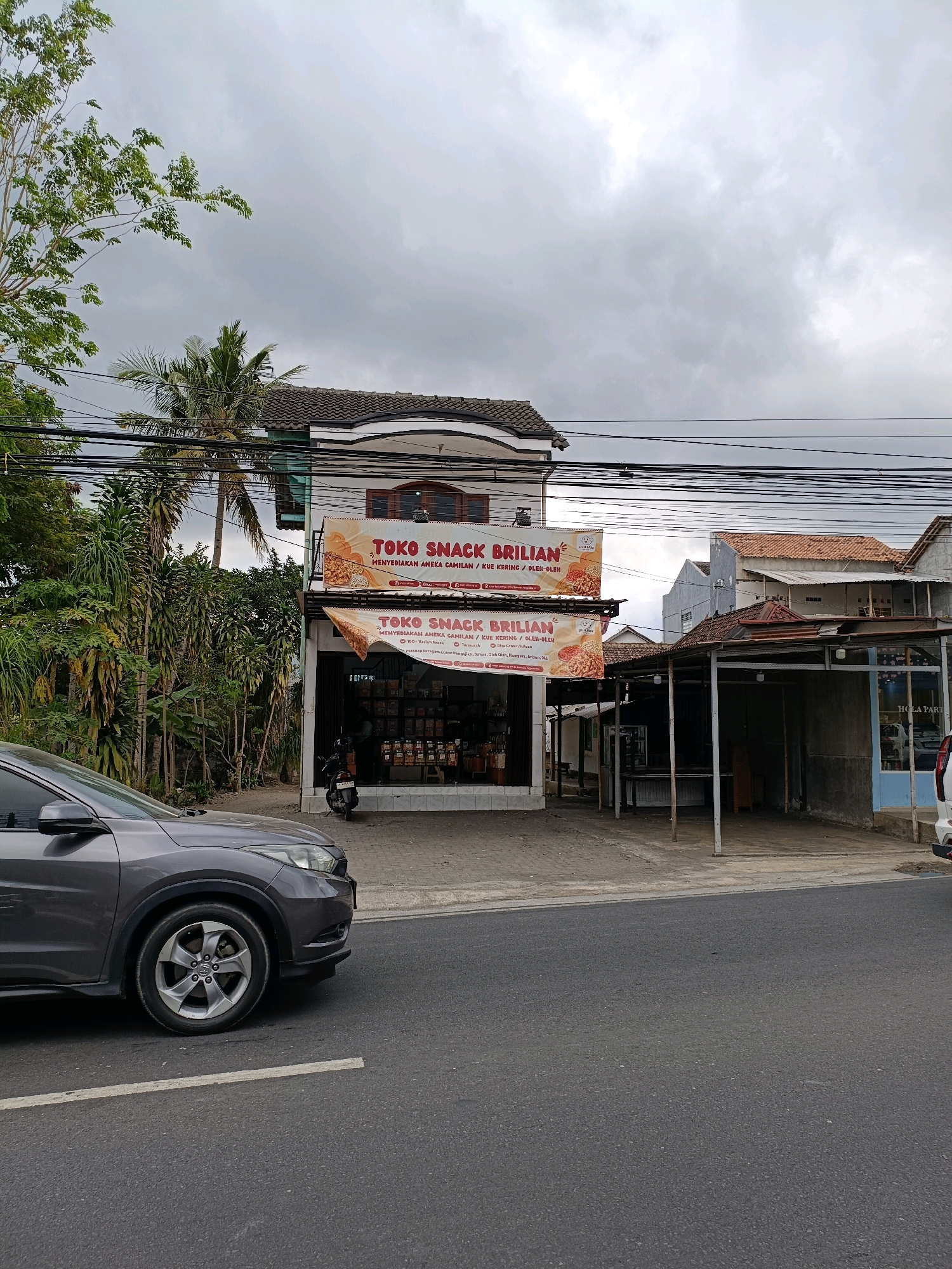 The image depicts a street scene in Ngaglik, Indonesia, featuring a storefront and a road with a car. The storefront, labeled "TOKO SNACK BRILLIAN," is a small retail establishment selling snacks and cookies. The building has two levels, with a prominent banner displaying the store's name and offerings. The lower level features an open entrance with shelves stocked with products, and a motorcycle is parked to the left of the entrance. The surrounding environment includes dense greenery, palm trees, and overhead electrical wires, indicative of a tropical or subtropical climate. The road in the foreground is asphalt, with a white line marking a lane division. A grey car is partially visible on the left side of the frame, positioned on the road. The sky is overcast, suggesting a cloudy day.