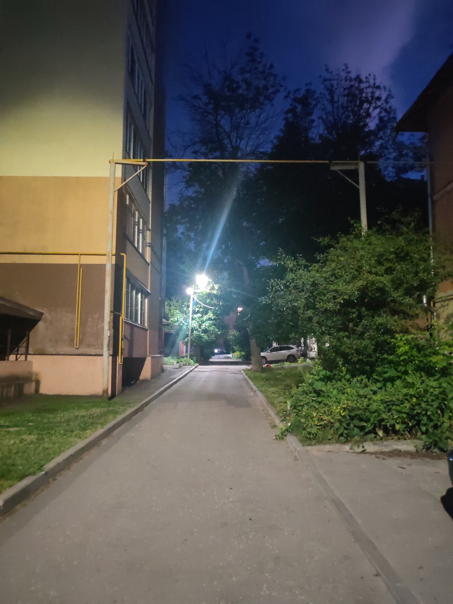 The image depicts a dimly lit alleyway in the city of Vladimir, Russia, at twilight or night. On the left, a portion of a multi-story building with light-colored walls and windows is visible. Yellow pipes run vertically along the side of this building. A sidewalk runs alongside the building, with a strip of grass and trees beyond it. The alleyway itself is paved, with a curb separating it from the grass.
In the center of the alley, a street lamp illuminates the scene, casting a bright light and creating glare. Several trees with dense foliage are present, some partially obscuring the view further down the alley. In the distance, a white car is parked on the right side of the alley. The sky above is dark blue with some wispy clouds. On the right side of the image, another building, darker in color, is partially visible, with dense green bushes in front of it. The overall atmosphere is calm and quiet, suggesting a residential area after dark.