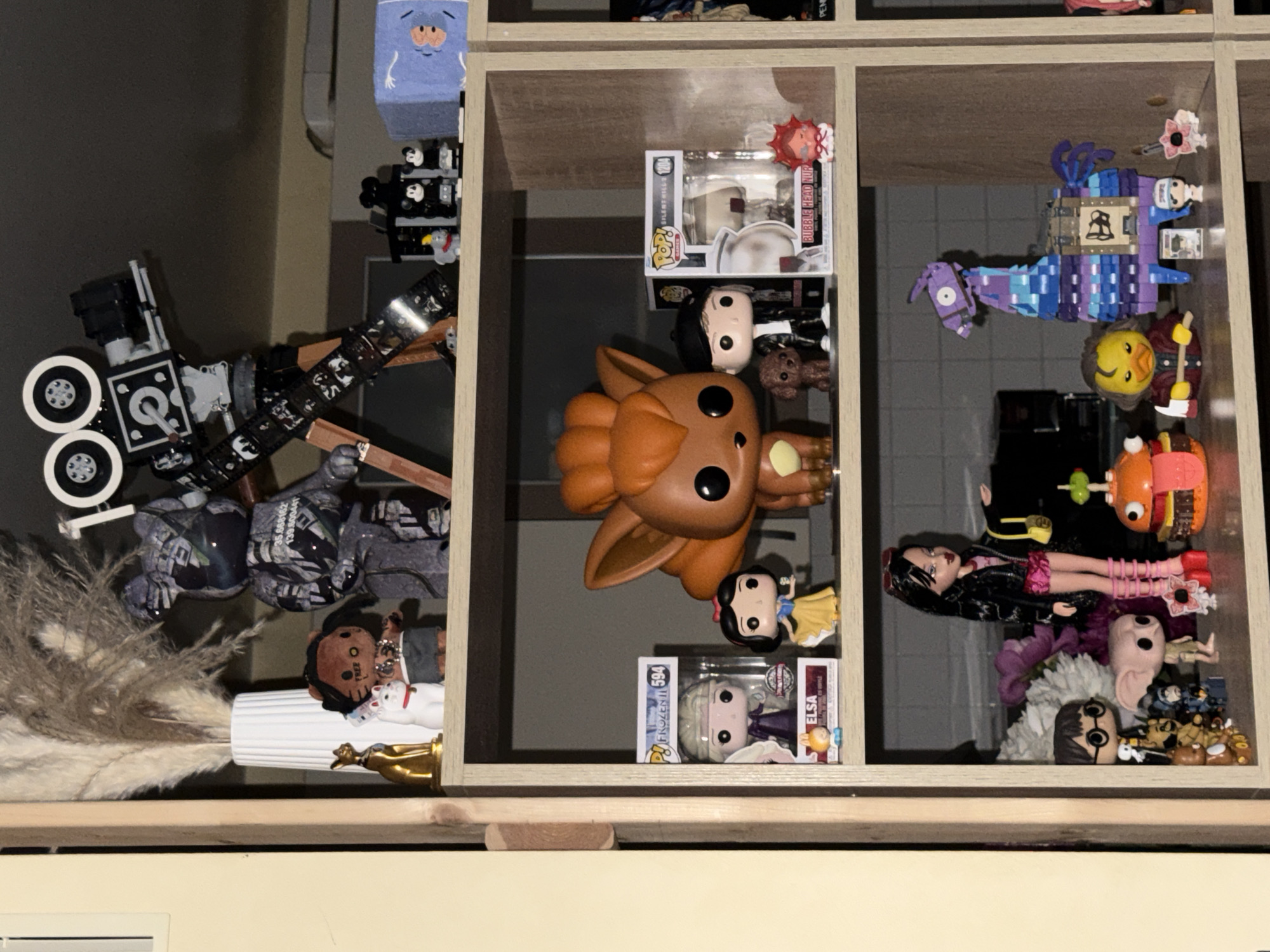 The image shows a wooden shelf unit containing various collectible figures and toys. The shelf is divided into three tiers.
The top tier displays a large grey and black patterned bear figure, a white vase with dried reeds, a small golden cat statue, a white cat figurine with a red collar, a doll with brown skin and dreadlocks, and a vintage-style movie camera made of LEGO bricks. To the right of the camera are two small Mickey Mouse figurines and a blue box with a white hand print.
The middle tier features several Funko Pop figures: Elsa from Frozen, Snow White, and a character with a black cap and a small brown dog. There is also a large orange and brown cartoonish fox-like creature. Further to the right is another Funko Pop figure in a box labeled "BUBBLE HEAD NUR," and a small red-haired figure.
The bottom tier contains a doll dressed in a black jacket and pink skirt, a Harry Potter Funko Pop figure, a Dobby figurine, a small teddy bear, a dark blue figurine, a burger-shaped toy, a yellow duck figurine holding an axe, a purple and blue LEGO llama, a small white Funko Pop figure, and a pink figurine with a white flower.
The background appears to be an indoor setting with a tiled wall. The overall impression is of a curated collection of pop culture memorabilia. There is no information in the image to indicate the location as Khimki, Russia.
